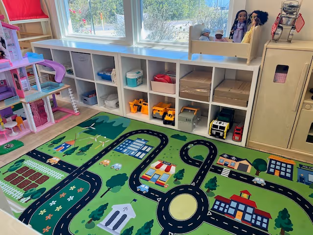 Colorful indoor playroom with cubby shelves, toys, dolls, and a road-themed rug.