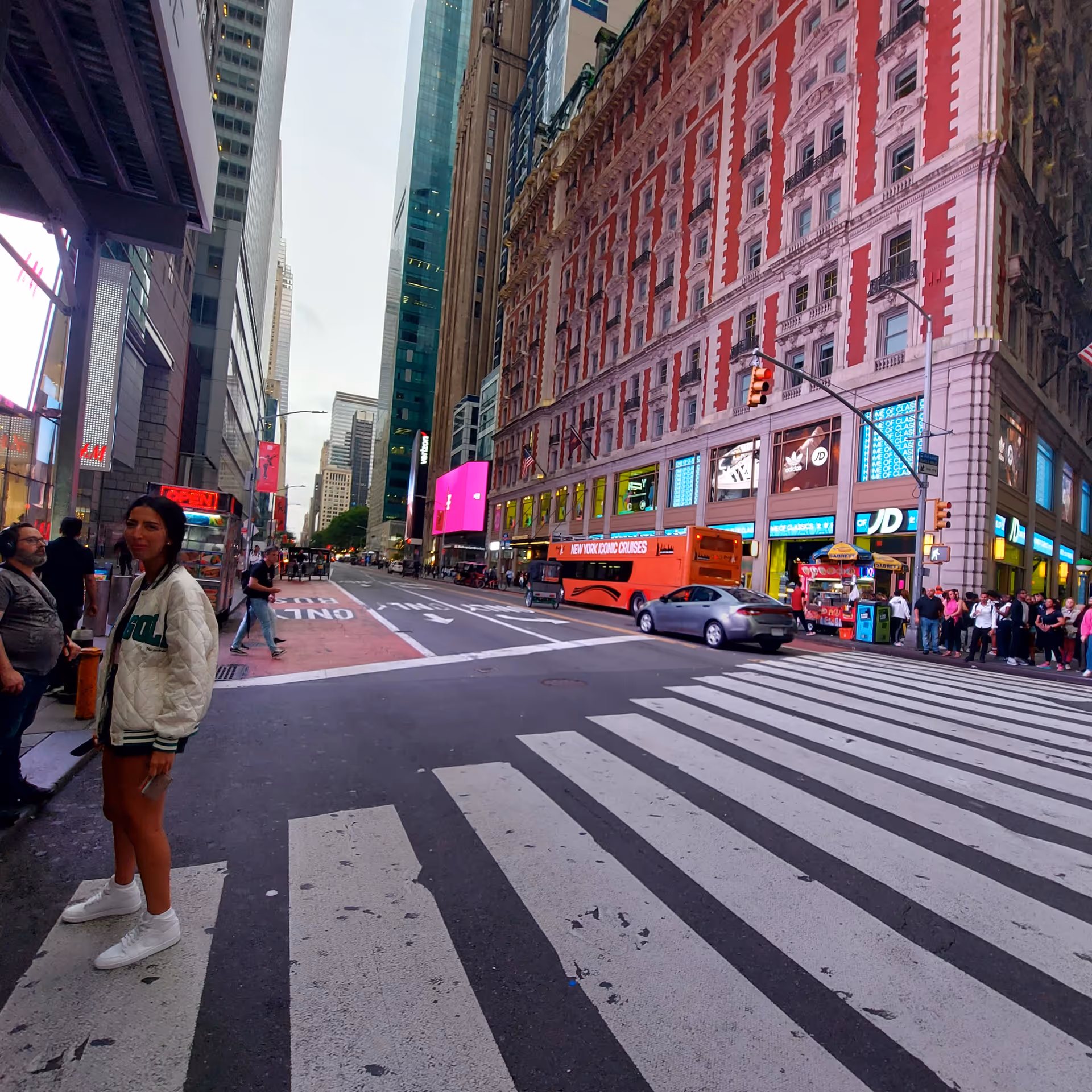 Busy city street with crosswalk, tall buildings, pedestrians, and an orange tour bus.