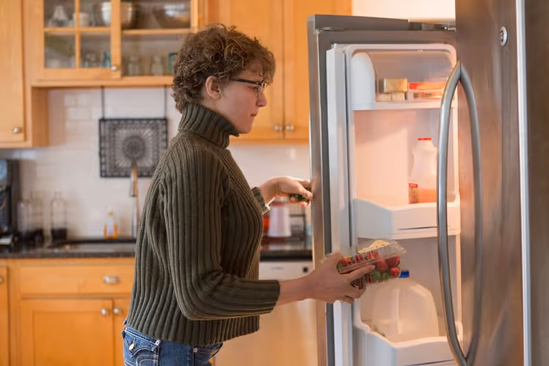 A person in a green turtleneck opens a fridge in a bright kitchen.