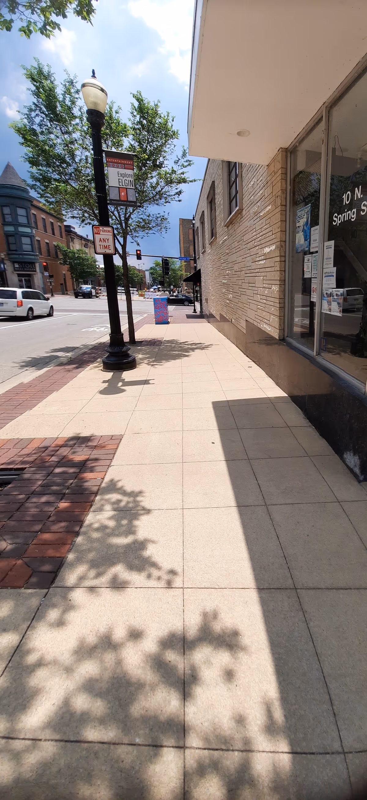 Sunny downtown sidewalk with storefronts, a streetlamp, and trees along the building exterior.