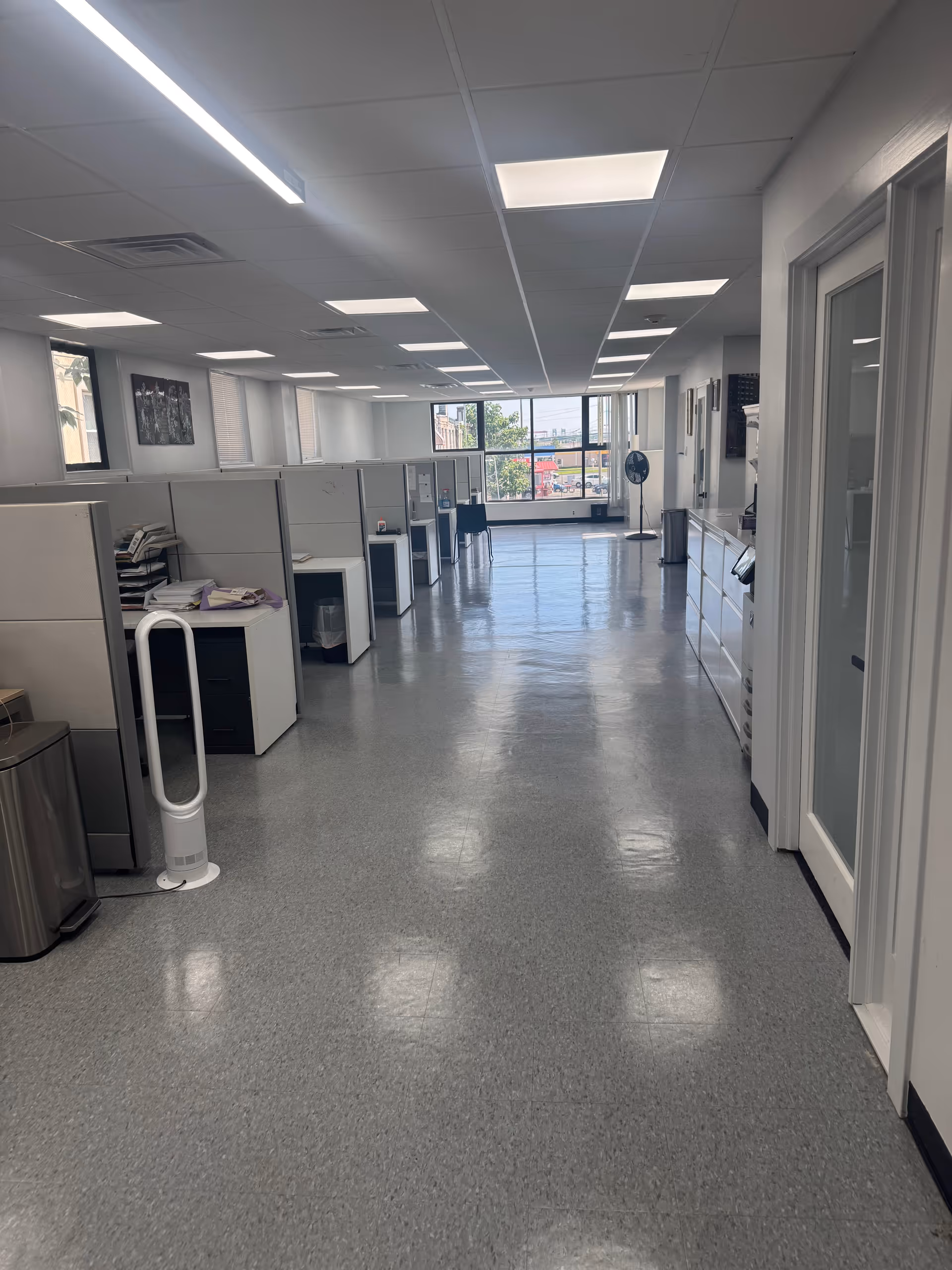 Long office corridor with cubicles, desks, and a standing fan near windows at the far end.