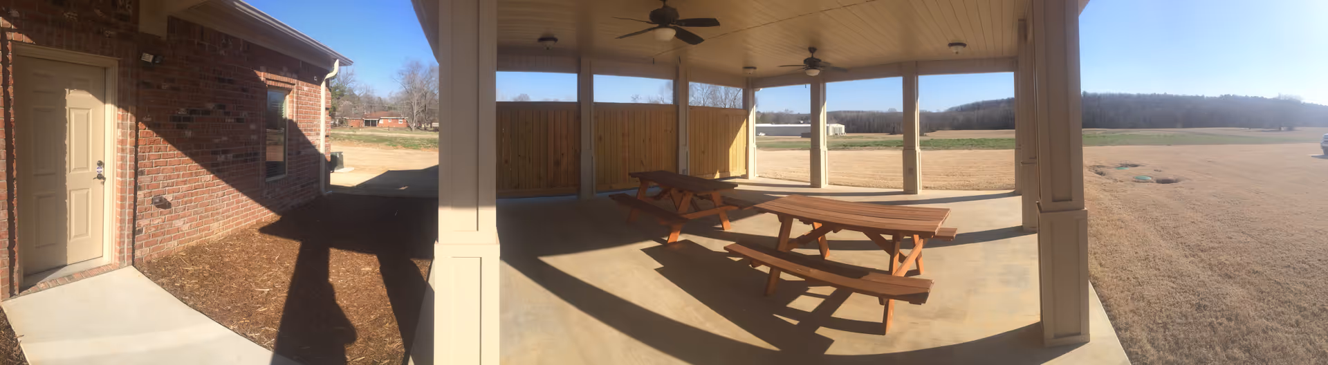 Covered outdoor patio with picnic tables, fans, and open lawn beyond.