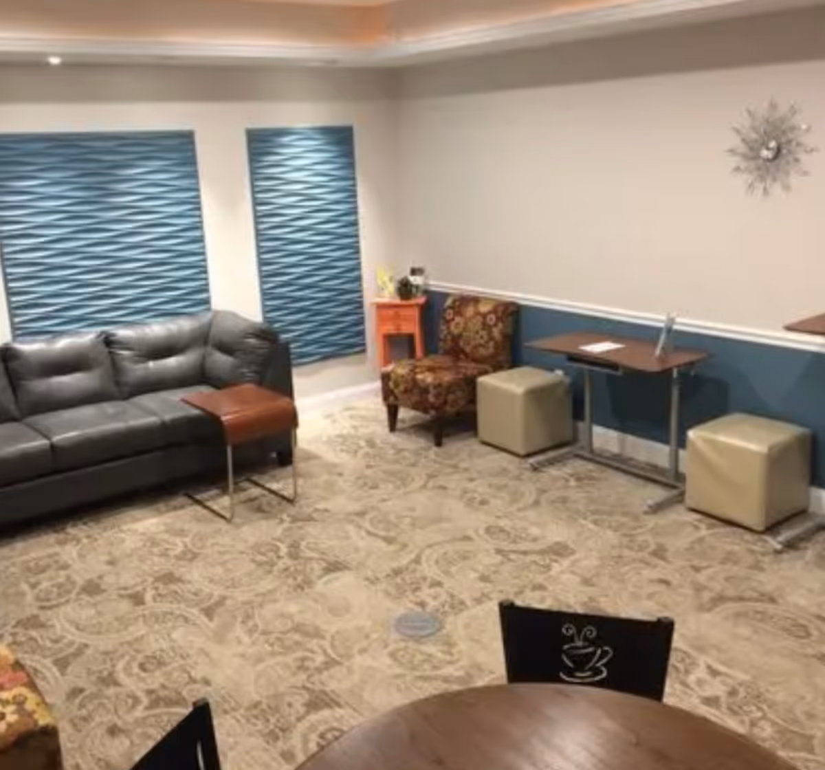 A cozy senior living lounge with blue blinds, sofa, chairs, and tables.