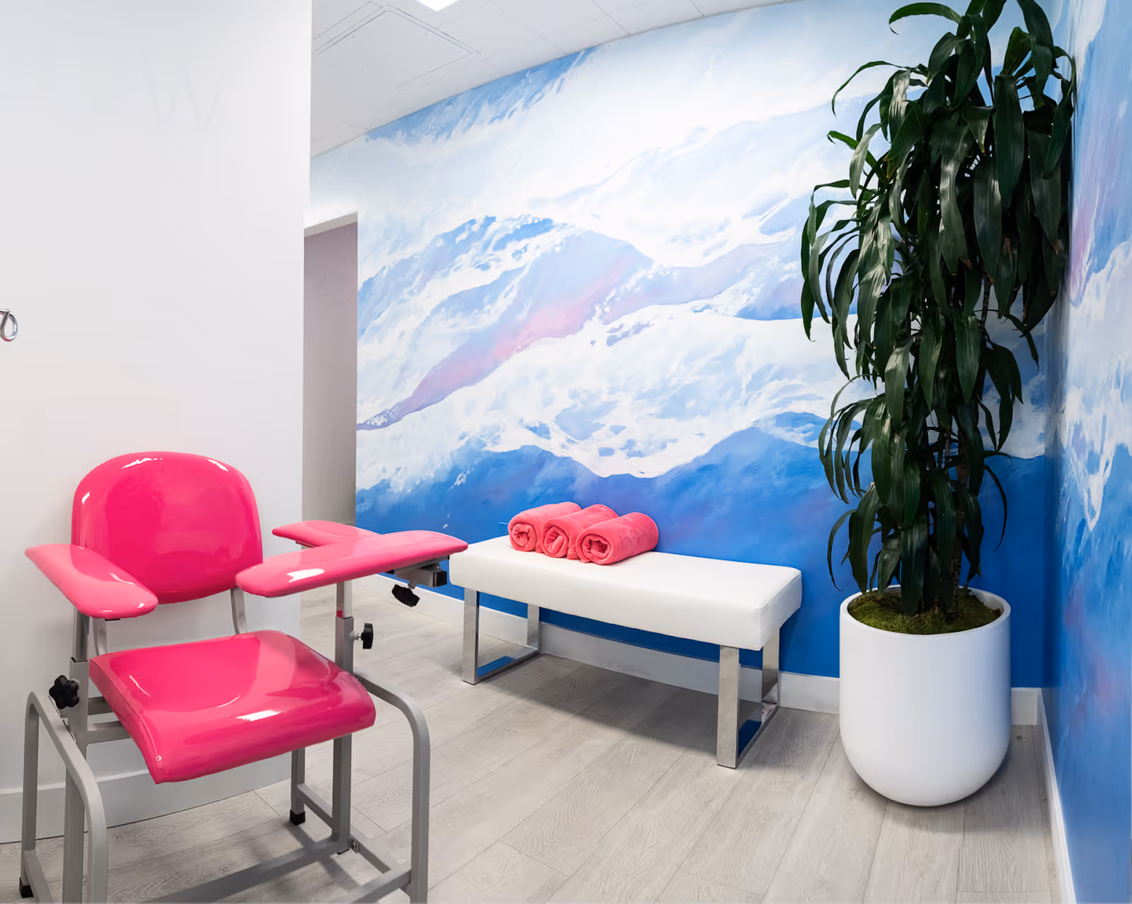Bright interior therapy room featuring a pink treatment chair, white bench, towels, and a potted plant.