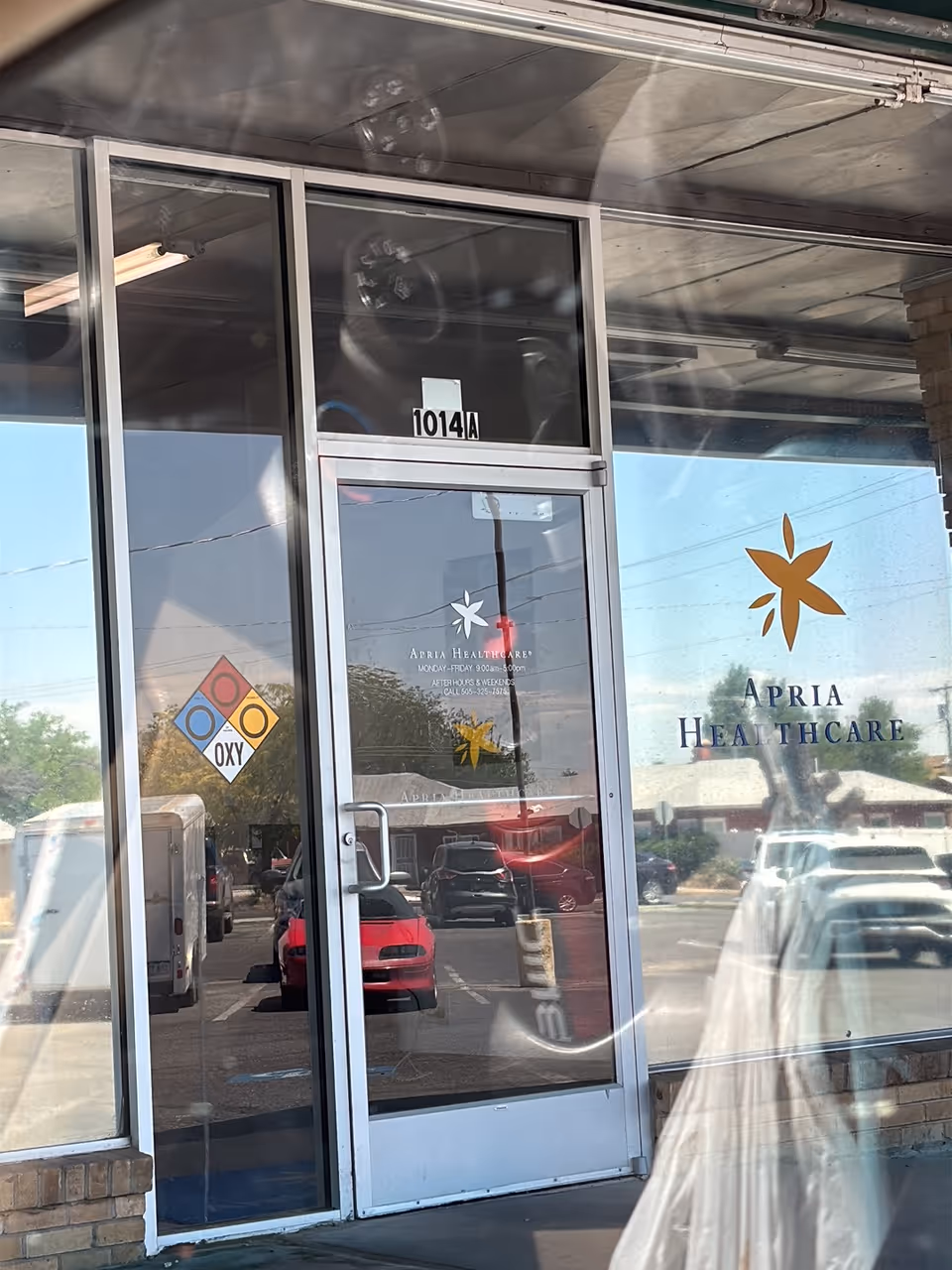 Glass storefront doors display Apria Healthcare signage with reflections of parked cars.