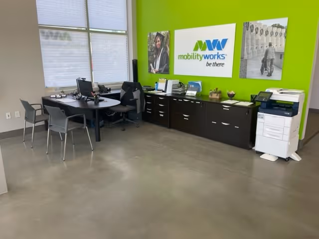 Office reception area with desks, chairs, filing cabinets, and a bright green wall displaying MobilityWorks logo.