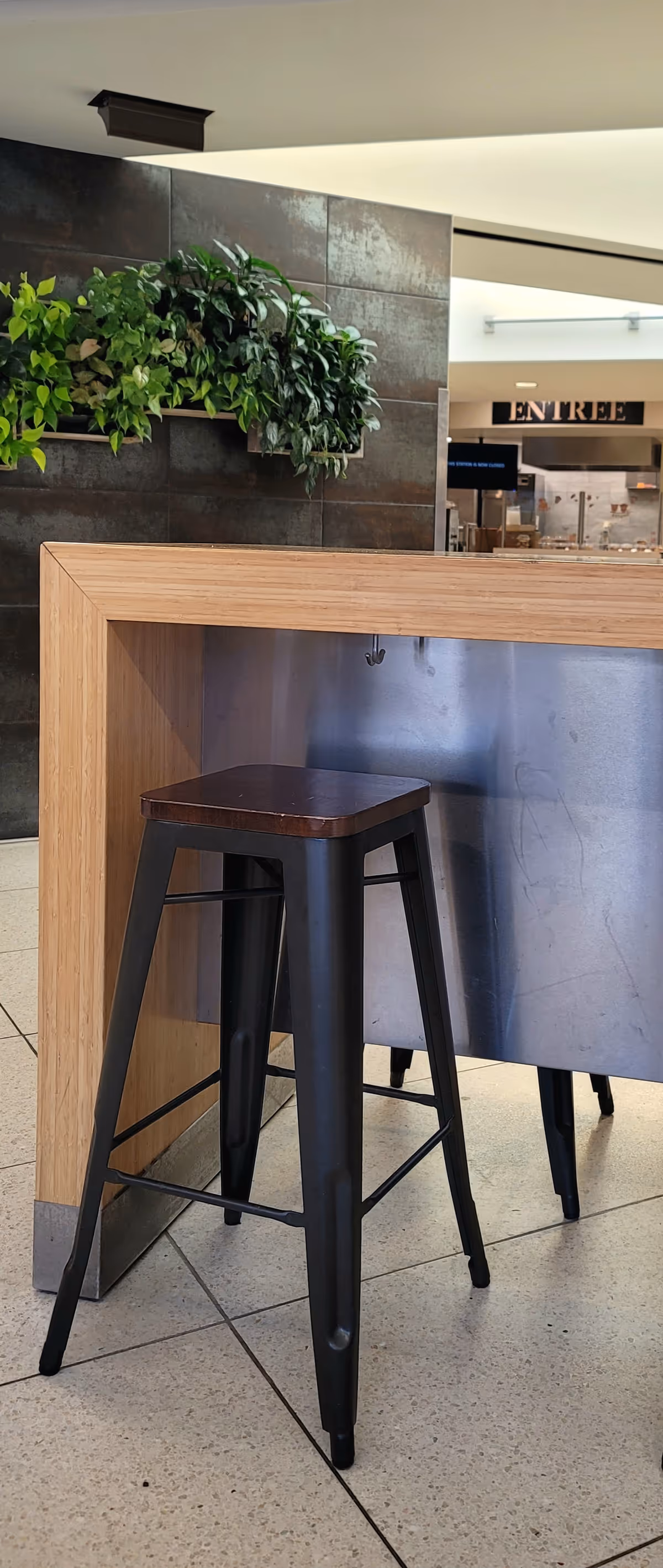 Indoor reception area with a wooden counter, metal panel, stool, and greenery.