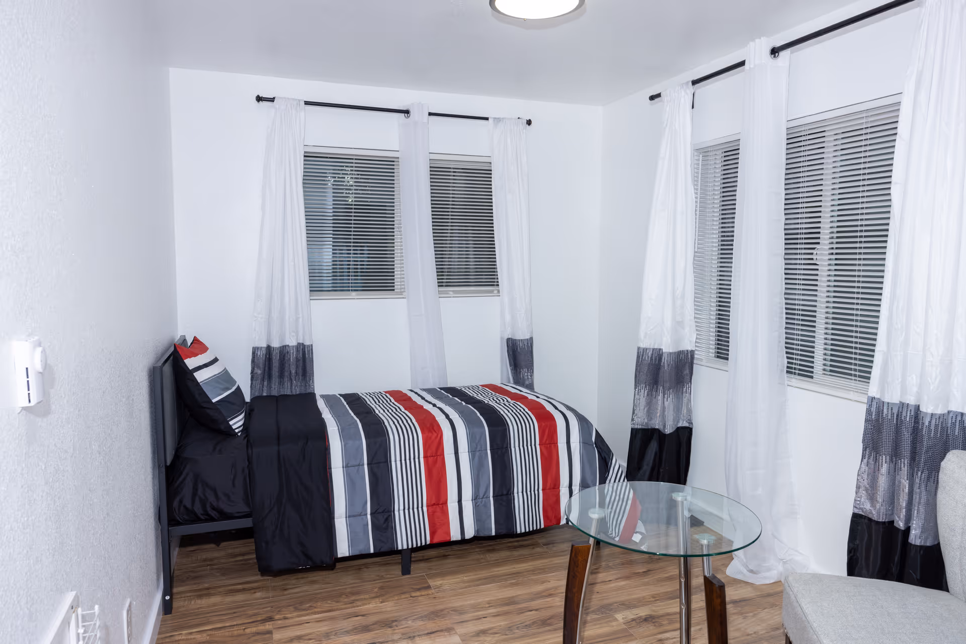 A compact bedroom featuring a striped comforter on a bed, white curtains, and a glass table.