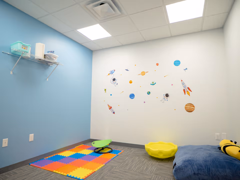Bright therapy playroom with space decals, blue wall, colorful foam mat, and a yellow bubble seat.