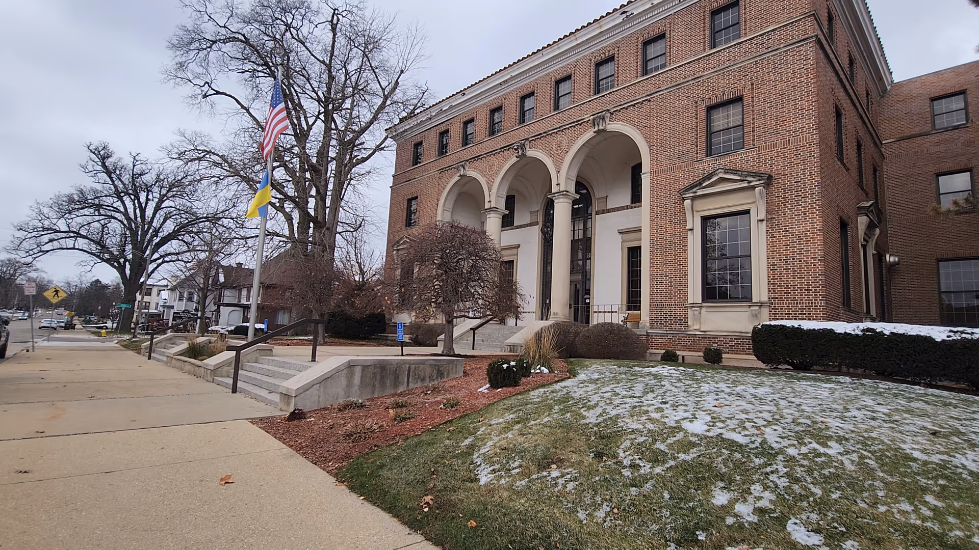 Brick building with arched front entrance, flagpoles, and a snow-dusted lawn beside a sidewalk.