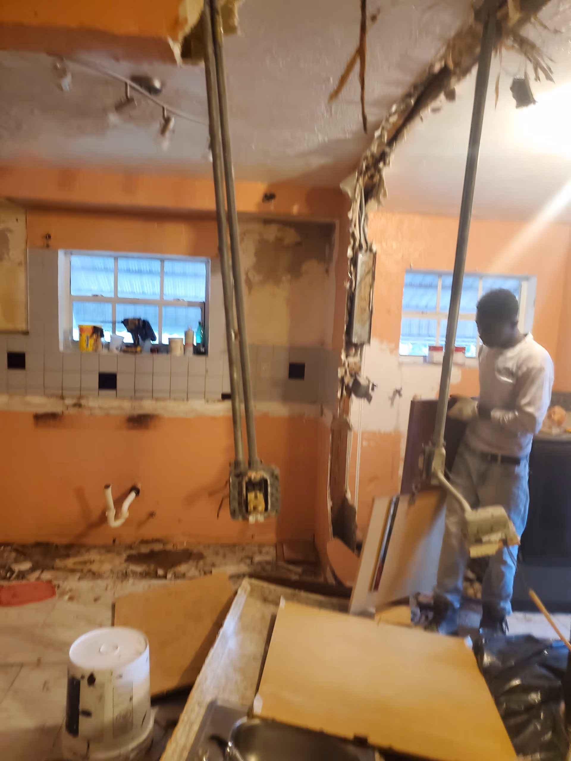 A kitchen undergoing renovation with exposed pipes, debris, and a worker inspecting equipment.