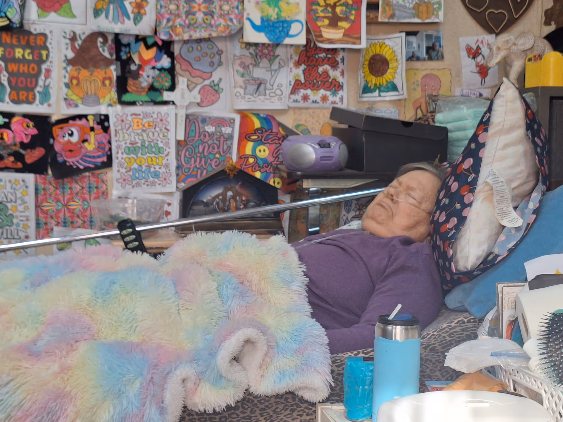 Elderly resident resting in bed amid colorful wall art and clutter.