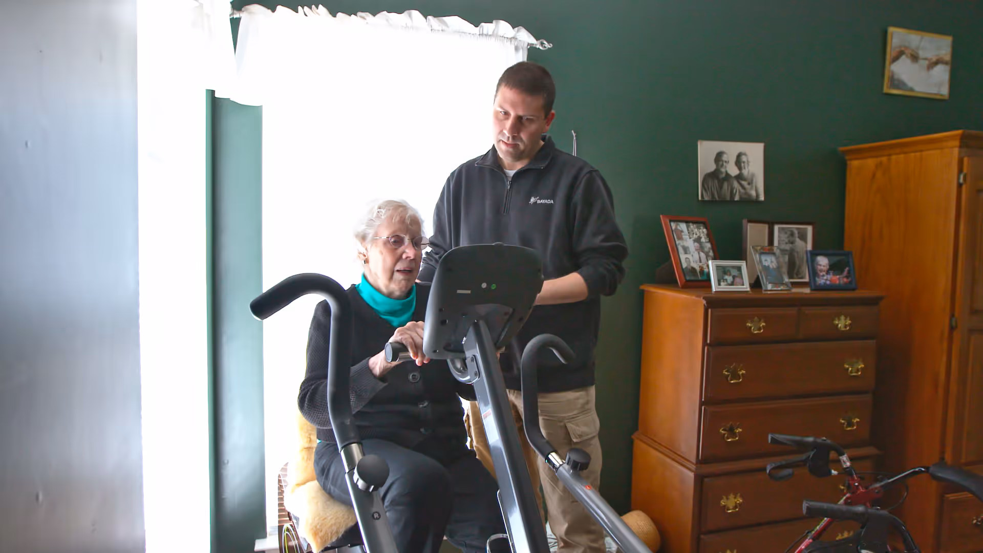 An elderly woman on a stationary bike assisted by a caregiver in a living room.