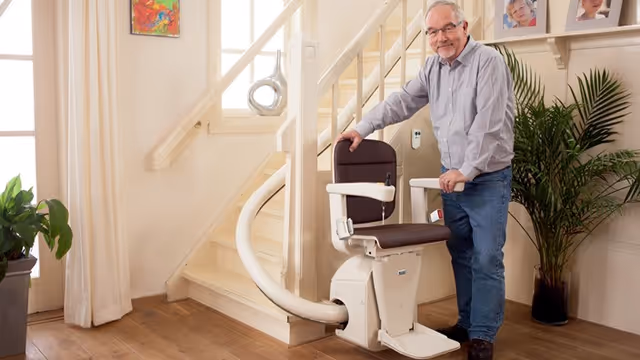 Older man stands beside a stair lift in a bright, homey facility interior.