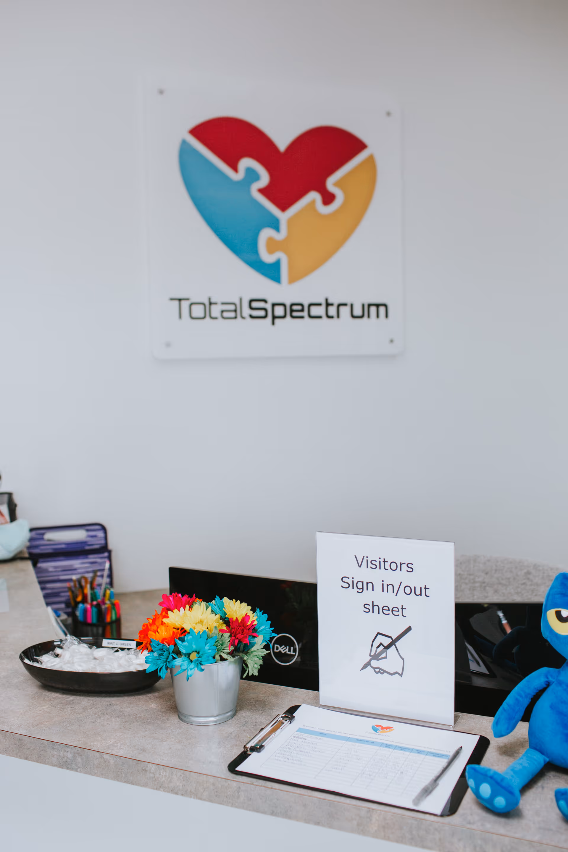 Reception desk with visitors sign-in/out sheet, colorful flowers, Total Spectrum sign, and a blue plush toy.