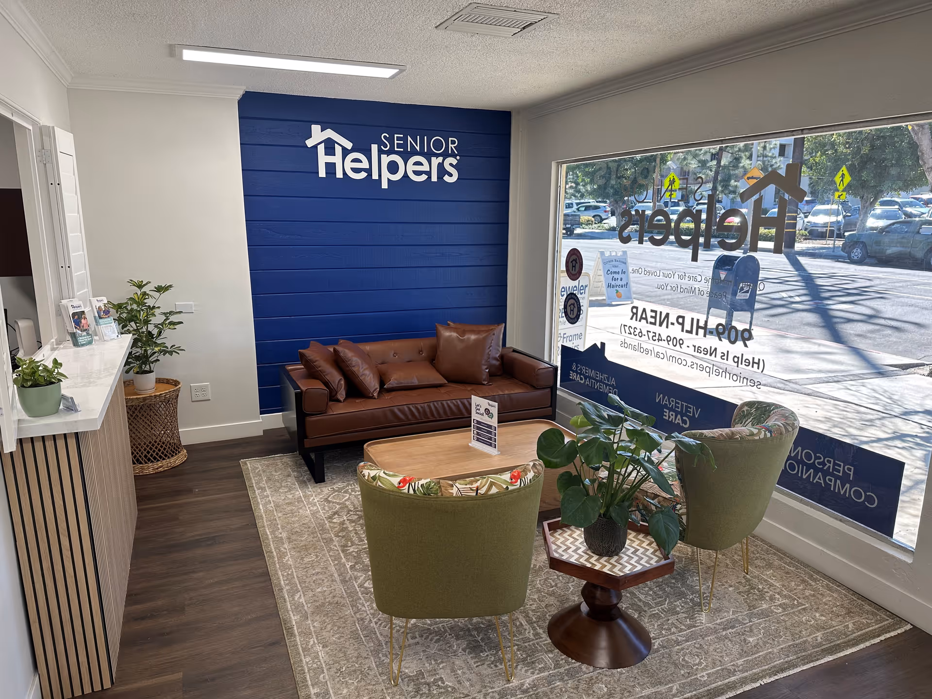 Reception lobby with blue wall Senior Helpers sign, brown sofa, green chairs, plants, and large window.