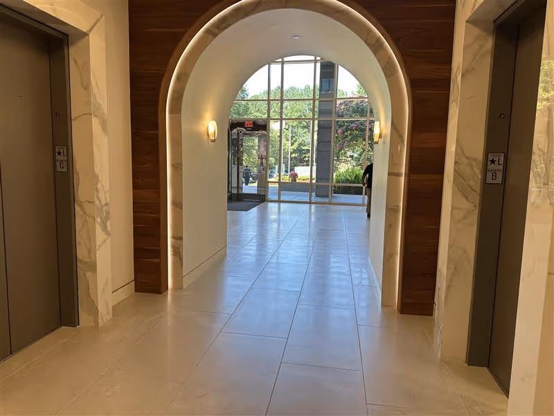 Bright marble hallway with arched entry, elevators on both sides, leading to glass doors outside.