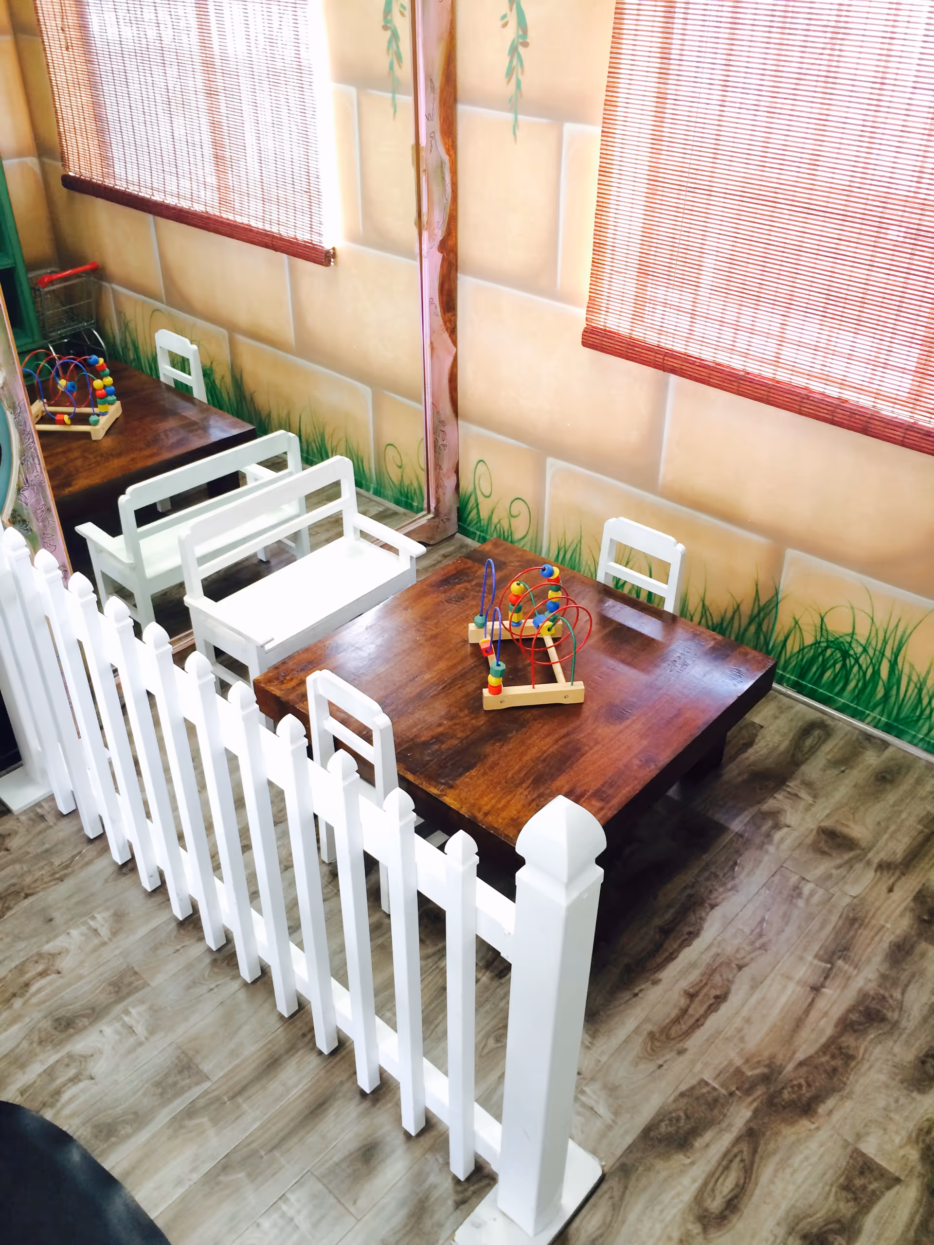 Indoor play area with white picket fence, wooden table, toys, and mural.