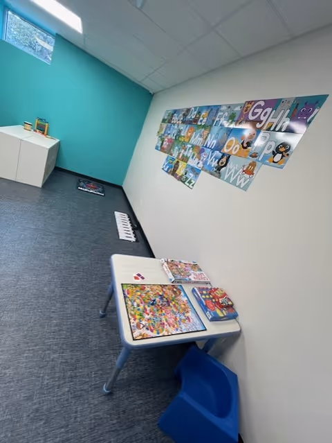 Small therapy room with a corner table, puzzles, teal wall, and alphabet mural.