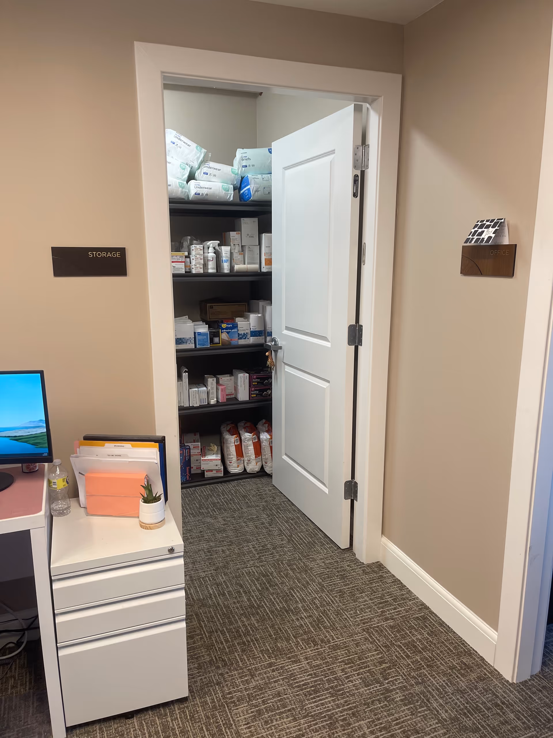 Office area with an open storage closet of medical supplies beside a desk.