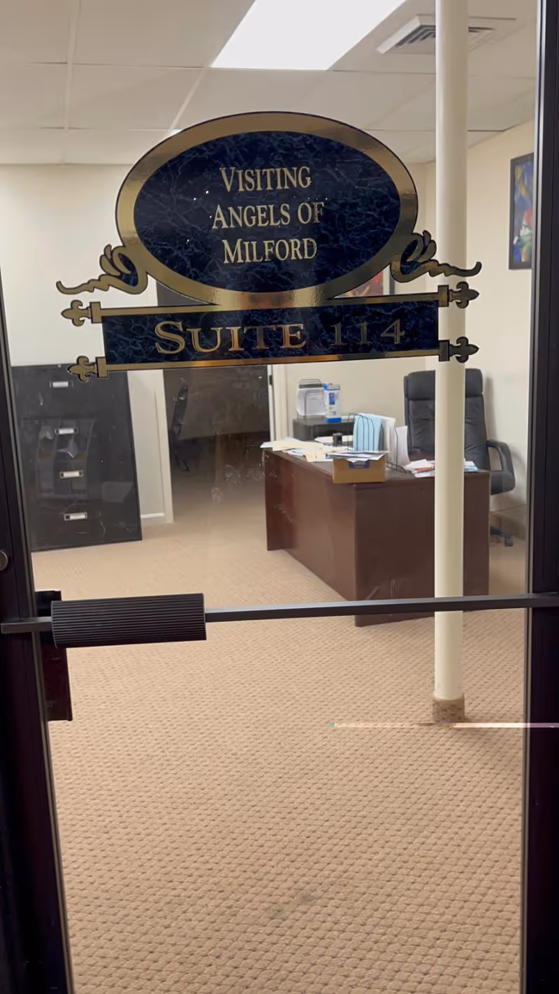 Glass door displays Visiting Angels of Milford Suite 114; office desk and chair visible inside.