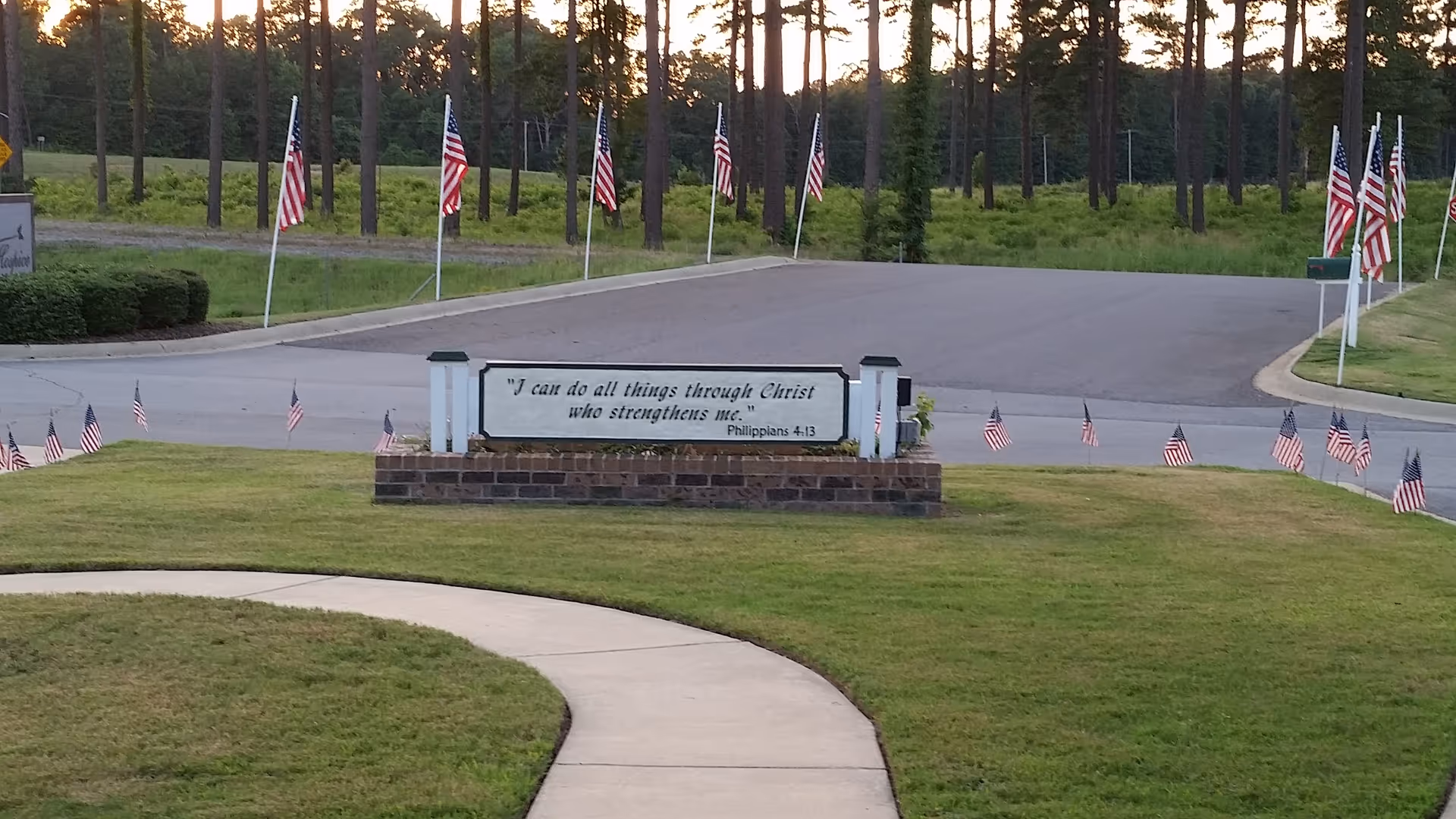 Grassy entrance with curved sidewalk, flag-lined road, and a brick sign displaying a scripture quote.