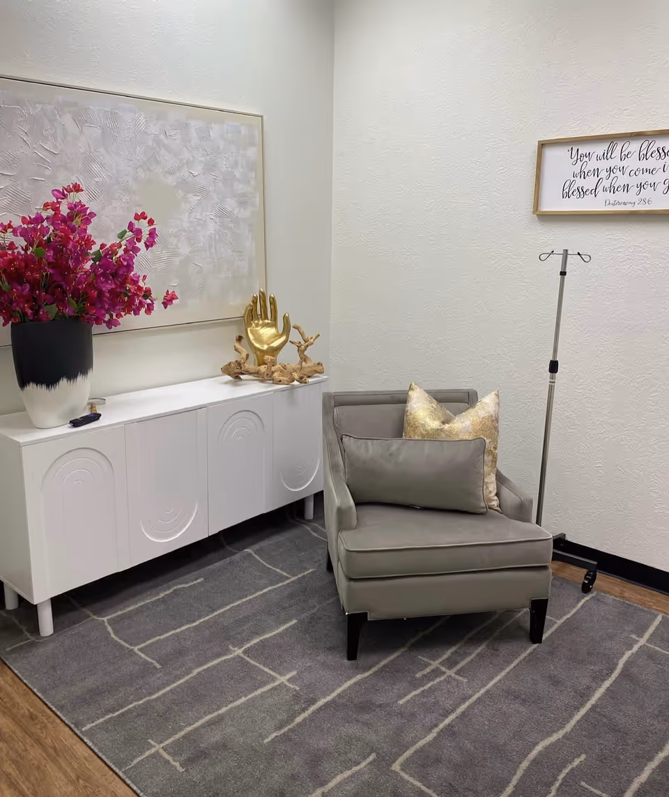 A cozy interior corner features a gray armchair, white cabinet, pink flowers, and wall art.