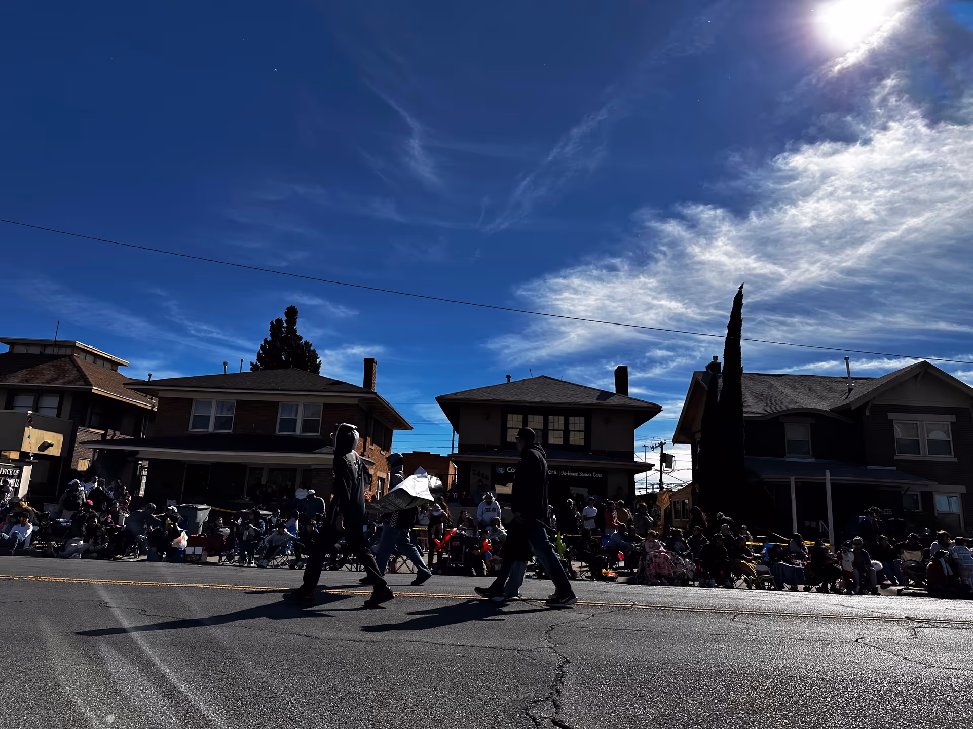 Sunny street scene with people walking and spectators seated along a row of houses.