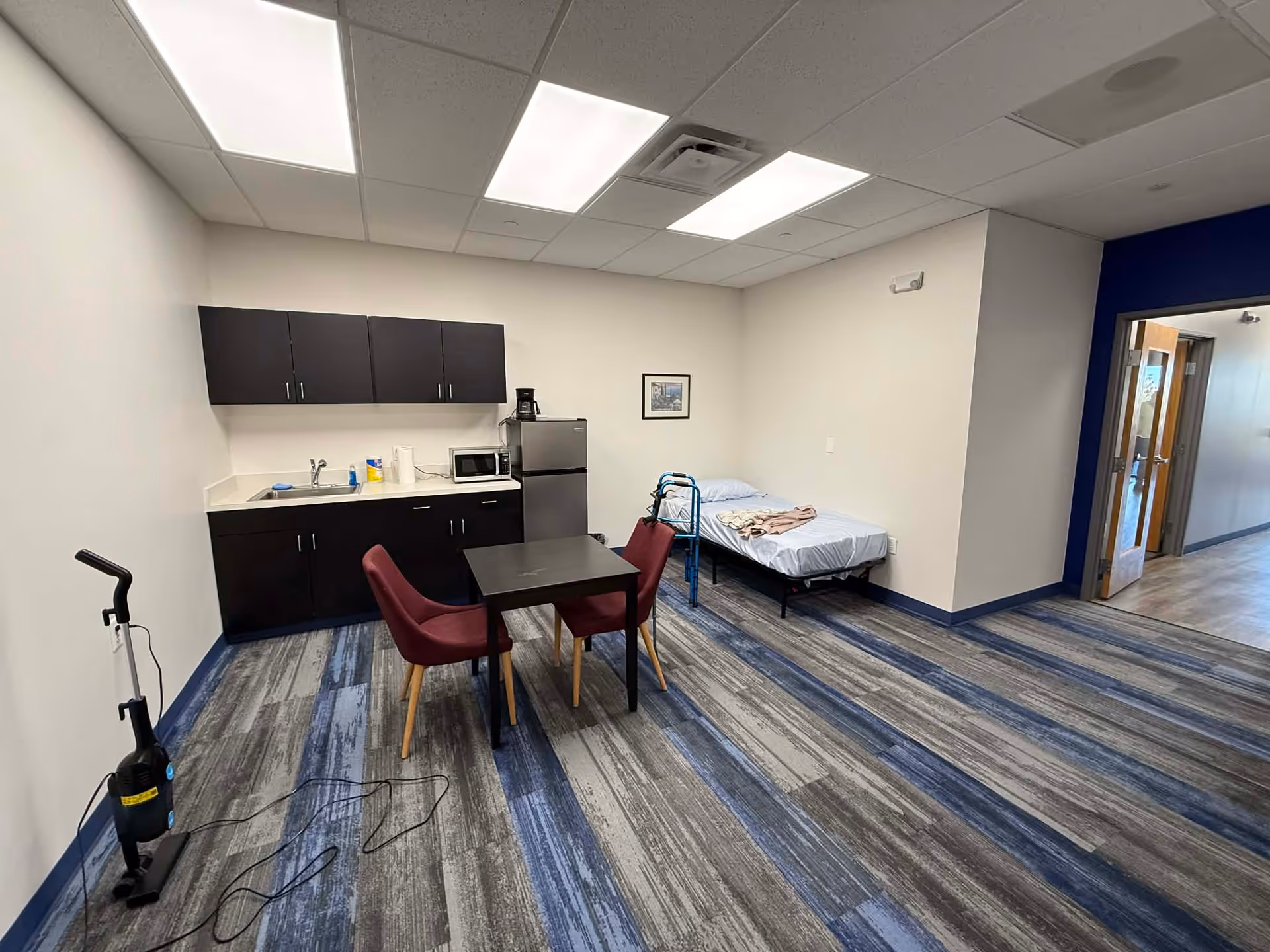 Interior senior living space with kitchenette, table and chairs, bed, and a vacuum on the floor.