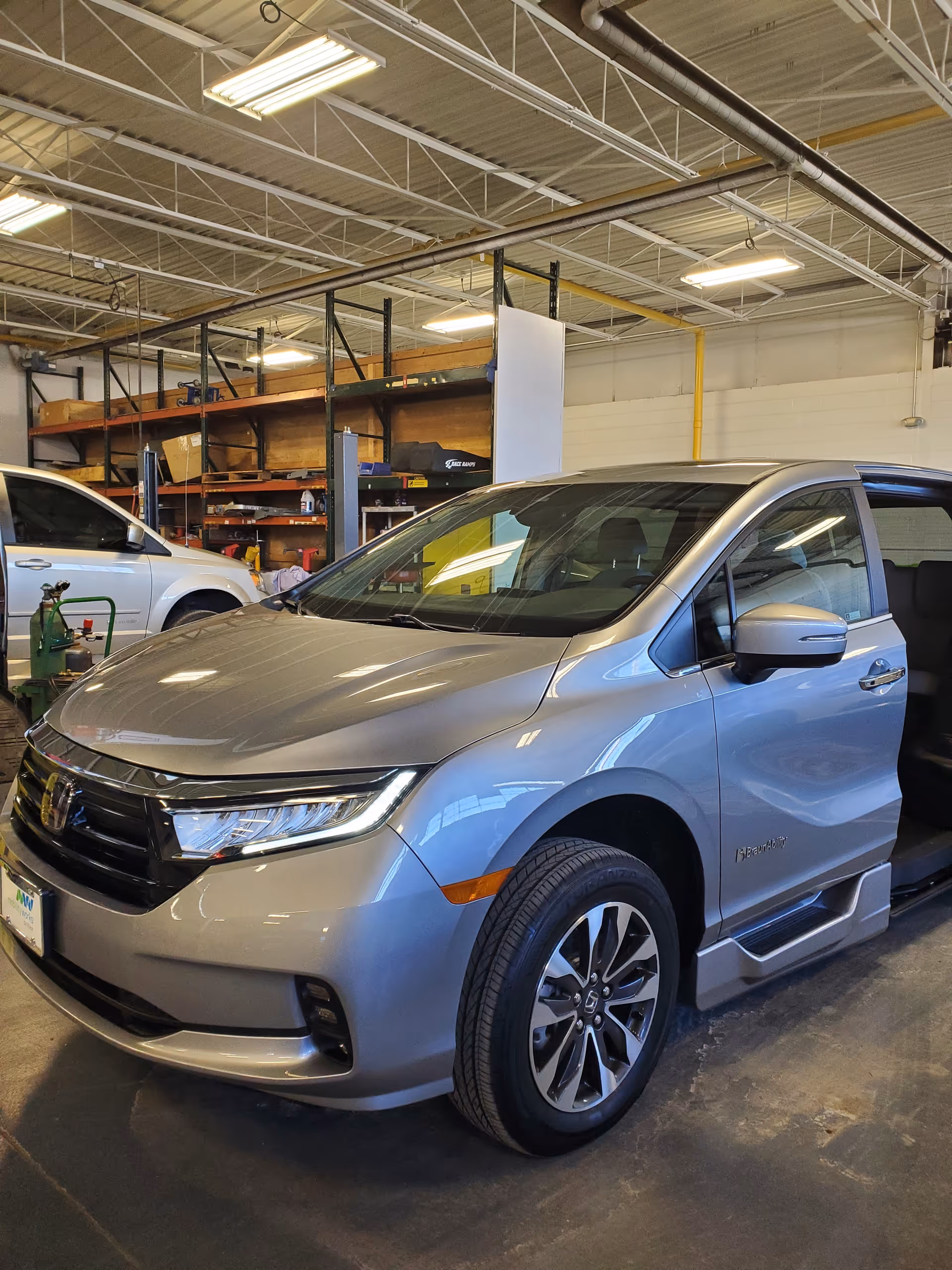 Silver minivan inside a warehouse-like repair shop with shelves and equipment.