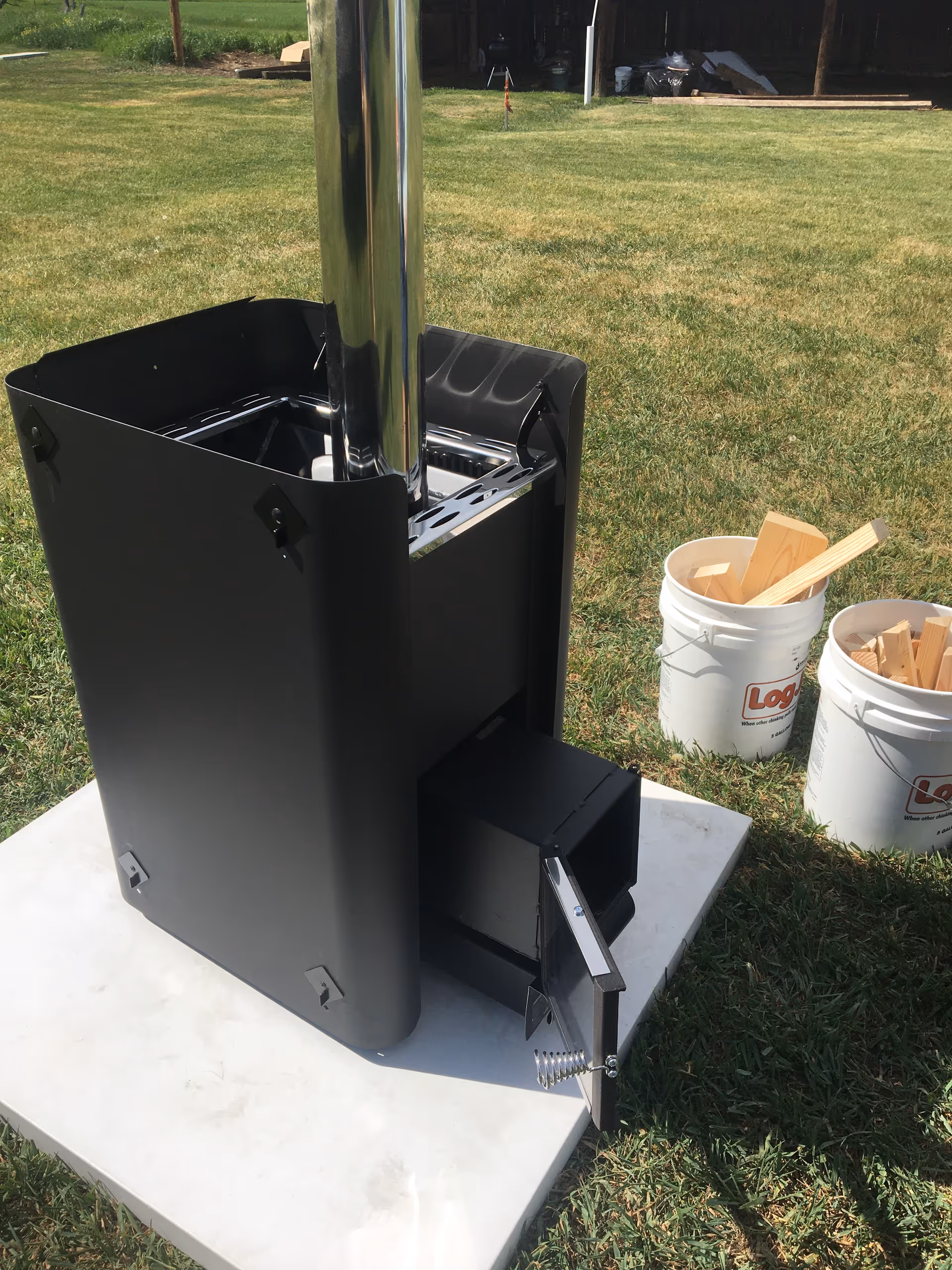 Black outdoor wood stove with chimney on a concrete pad beside wood buckets.