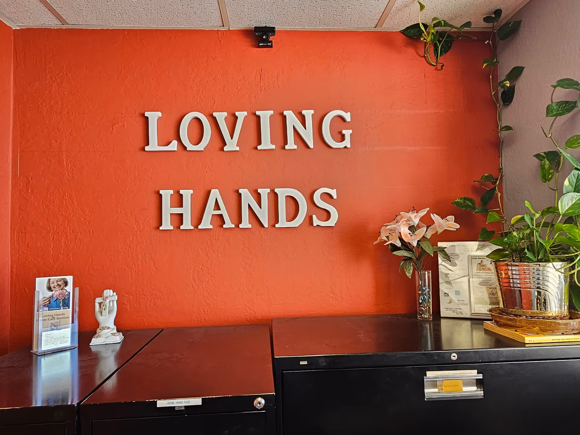 Red interior wall with large LOVING HANDS sign and potted plants in a reception area.