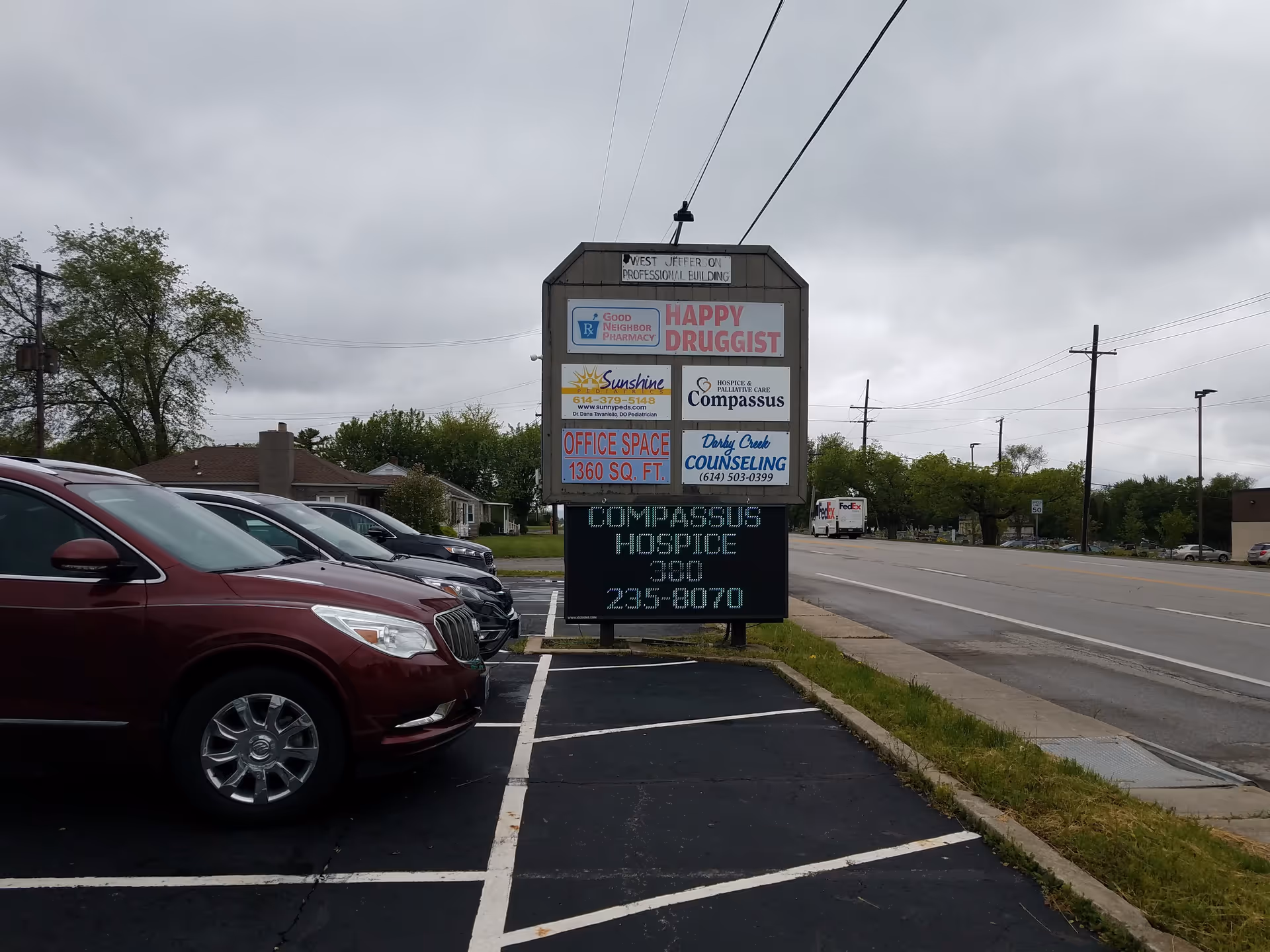 A roadside signboard advertising Compassus Hospice beside a parking lot under cloudy sky