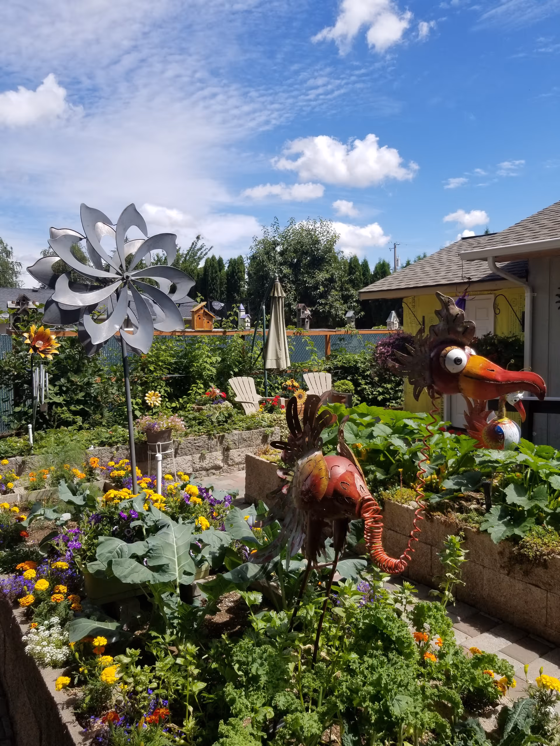 Colorful garden with flowers, wind sculptures, and a house exterior in a sunny yard.