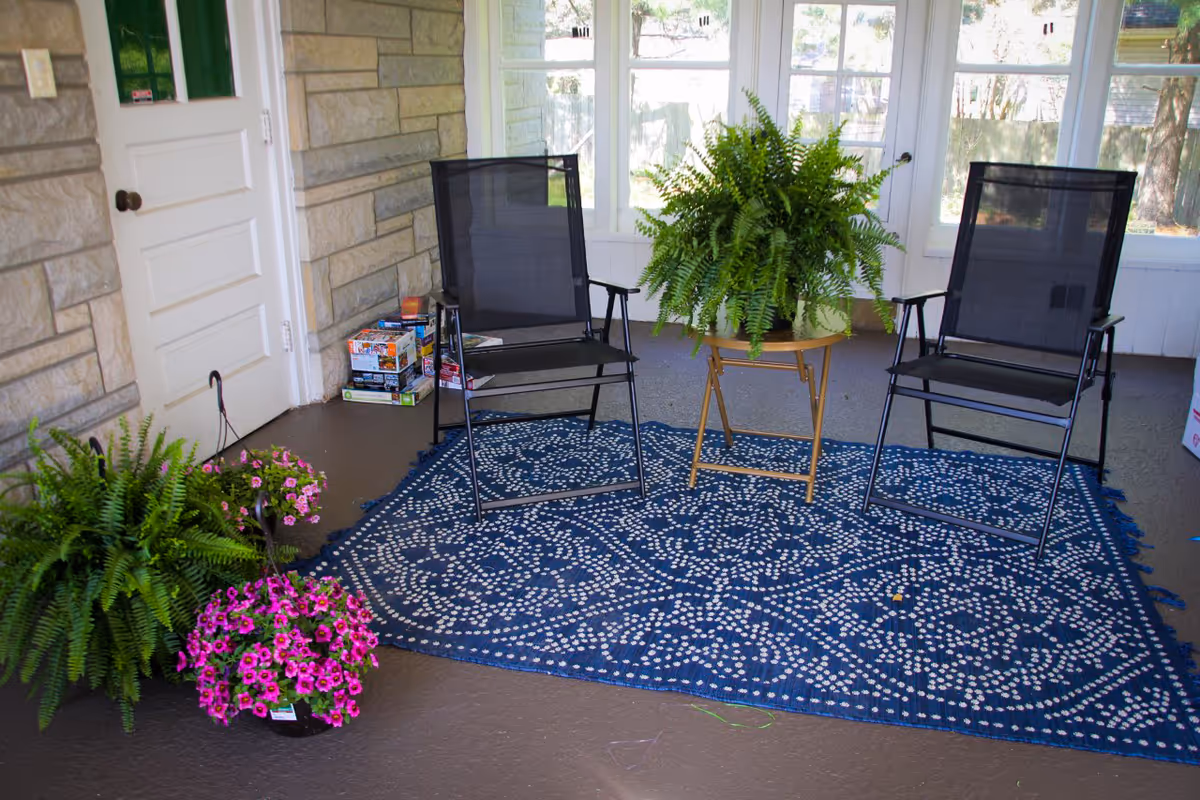 Covered porch seating area with two chairs, a fern centerpiece, potted flowers, and a patterned rug.