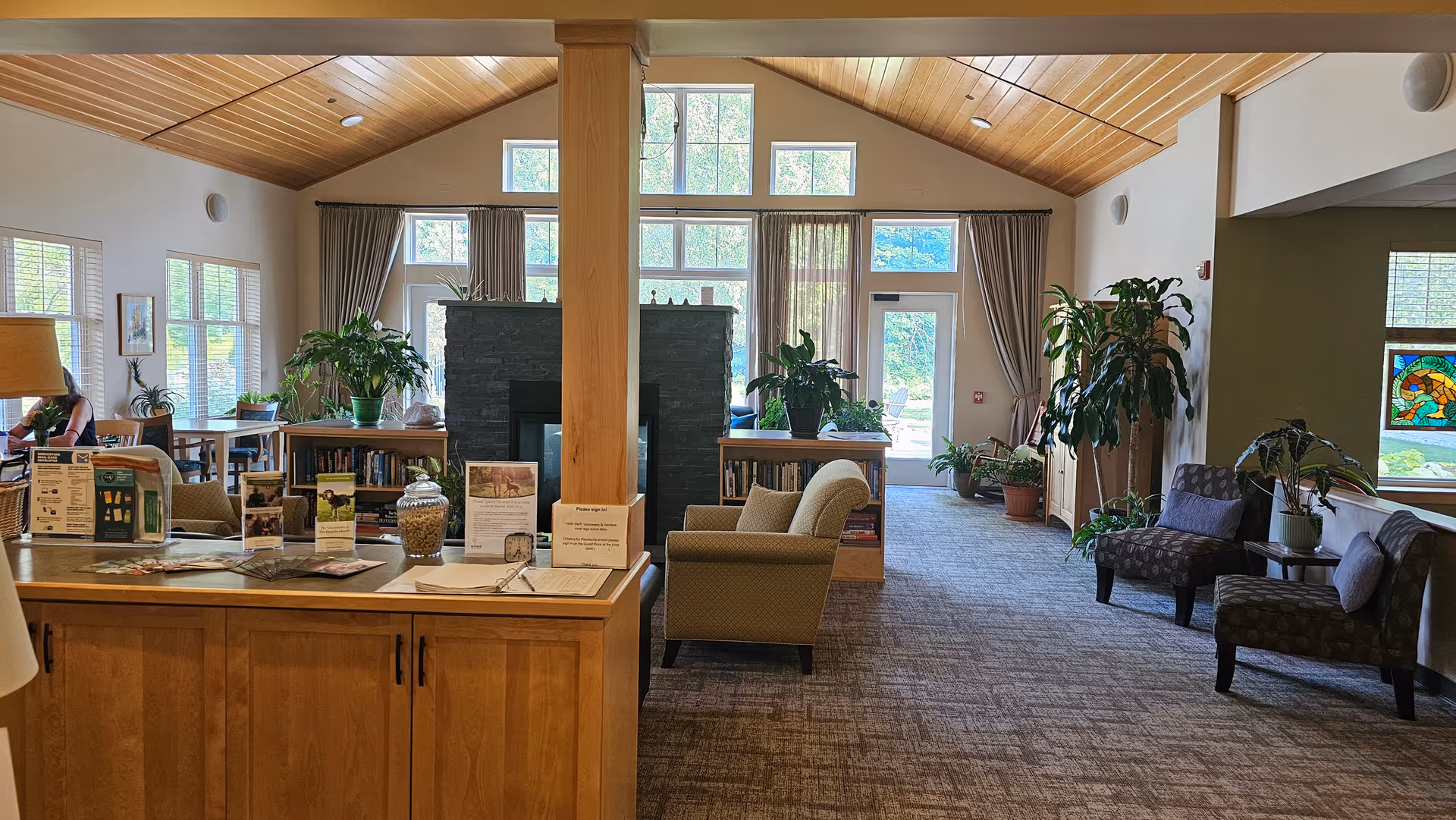 Spacious senior living lounge with wood-beamed ceiling, fireplace, plants, and seating.