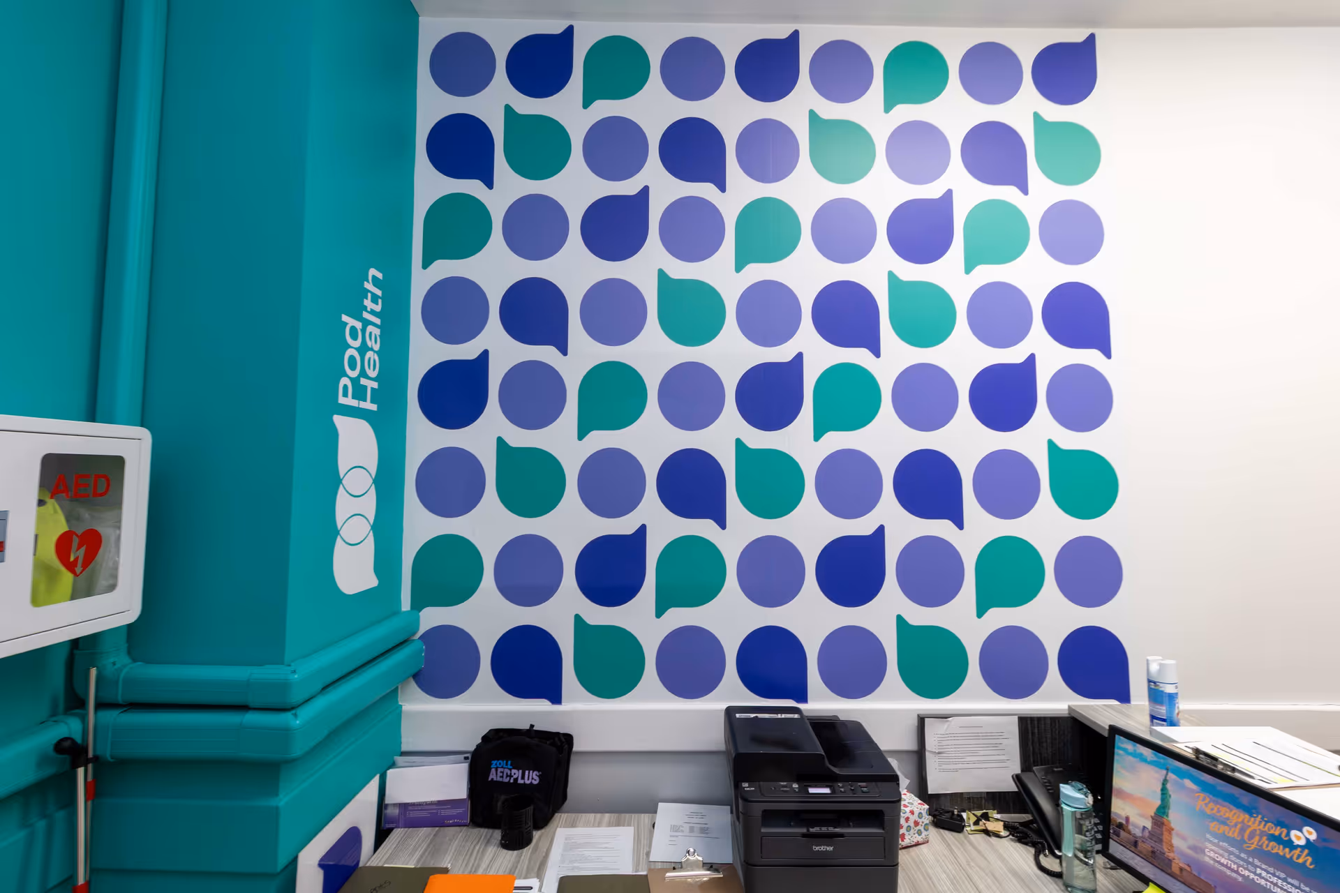 Teal wall with geometric teal-purple wallpaper and an office desk with printer and AED cabinet.