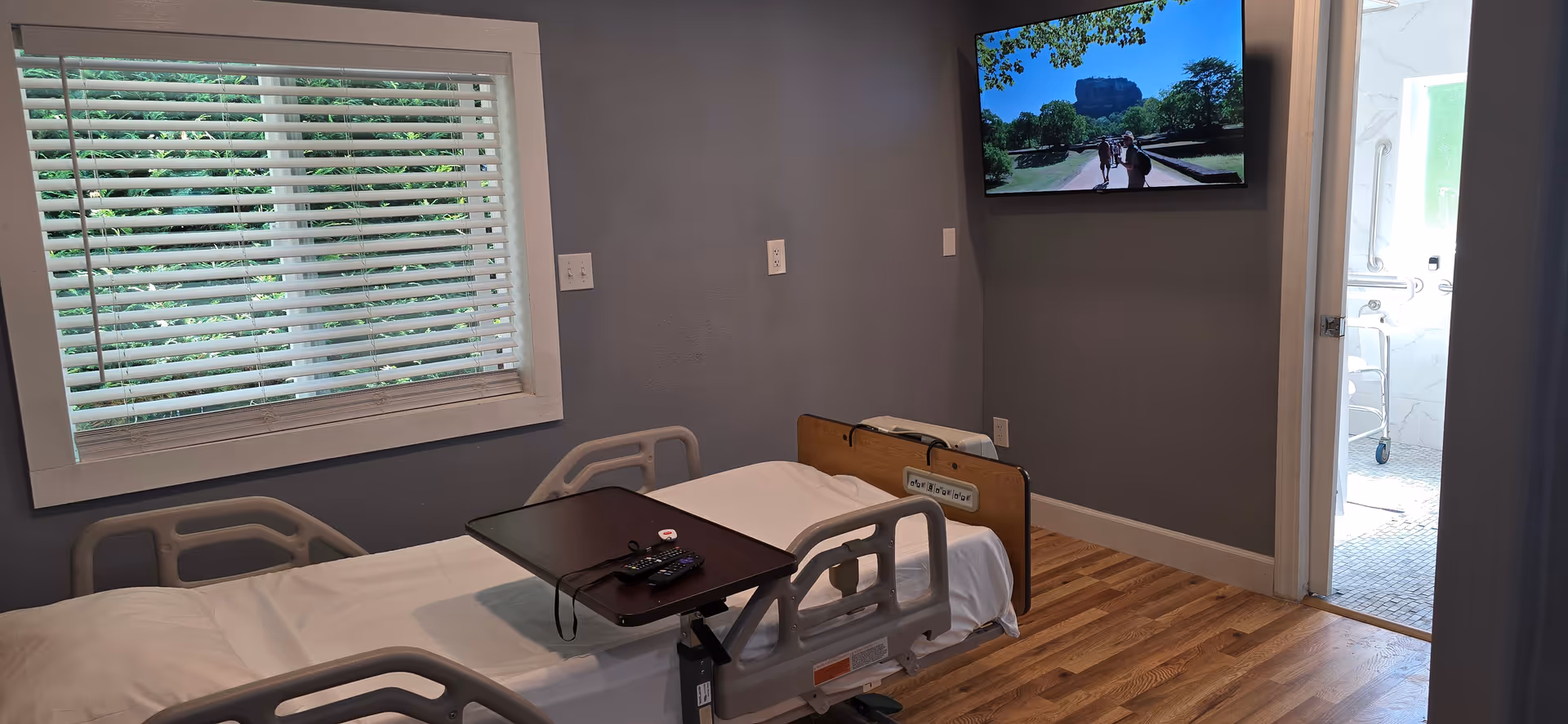 Bedroom-like hospital room with a bed, window blinds, TV, and doorway to a bathroom.