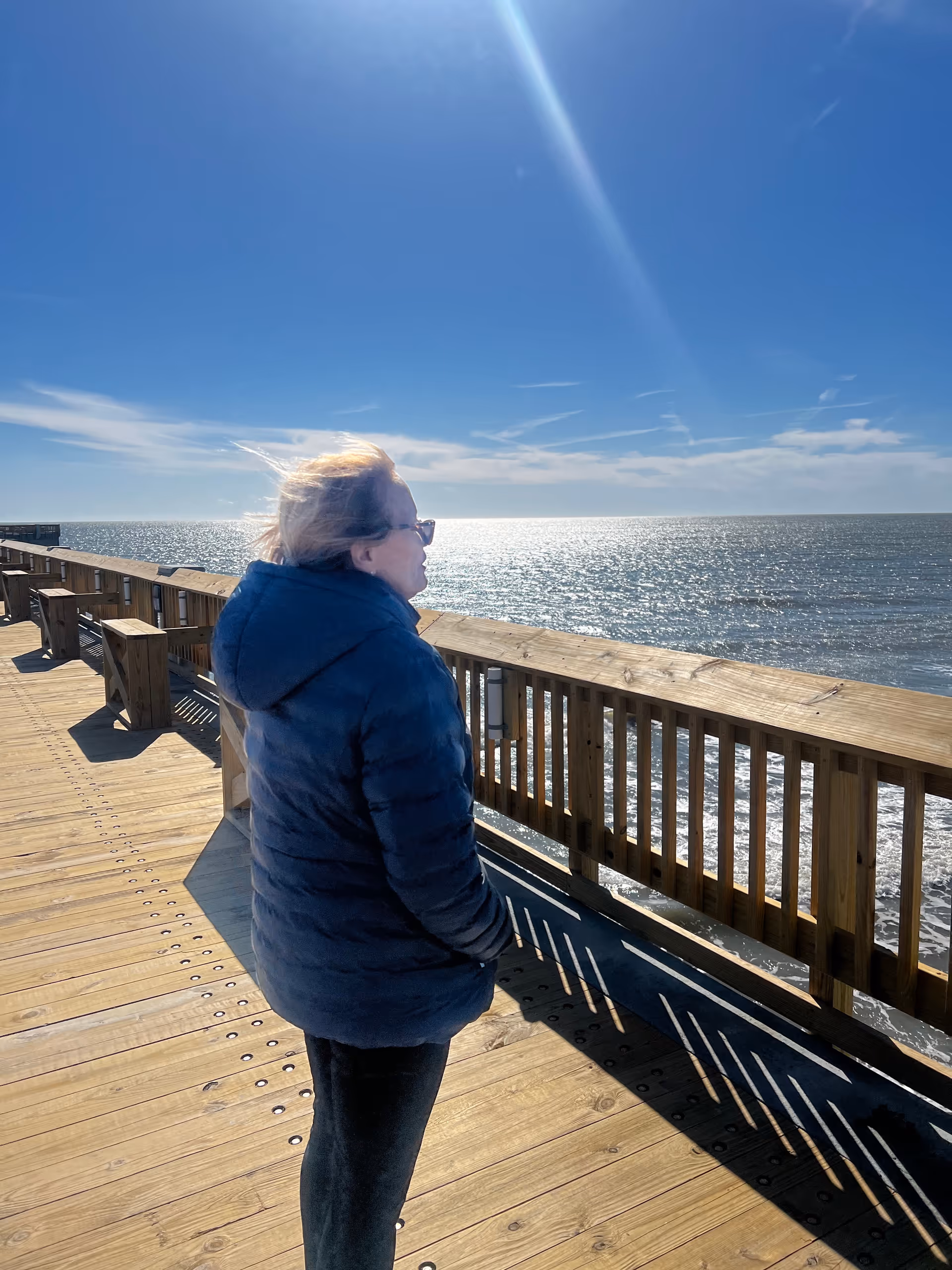 A person in a blue puffer jacket stands on a wooden boardwalk overlooking the ocean.