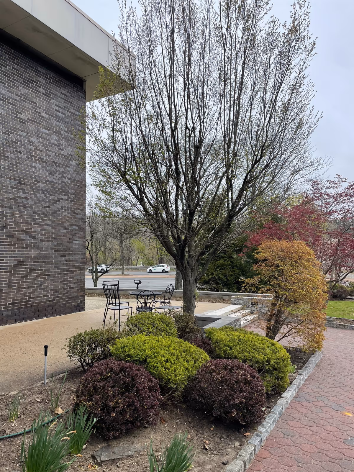Outdoor seating area with metal table and chairs beside a brick building and landscaped shrubs.