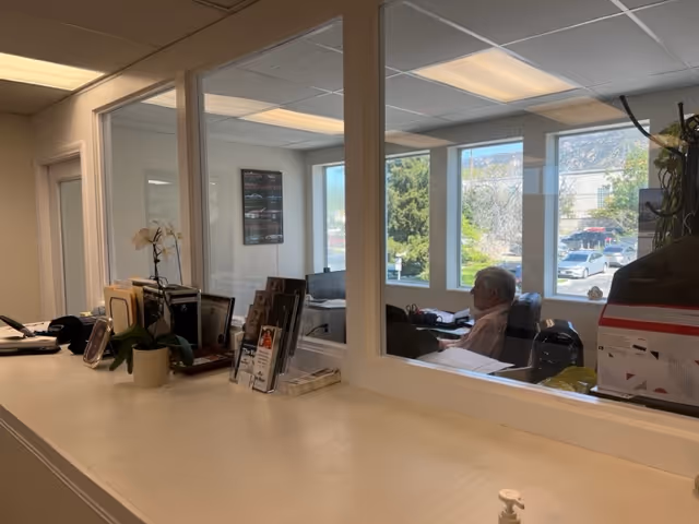 Reception desk with brochures and a plant, seen through a glass partition; a man sits nearby.