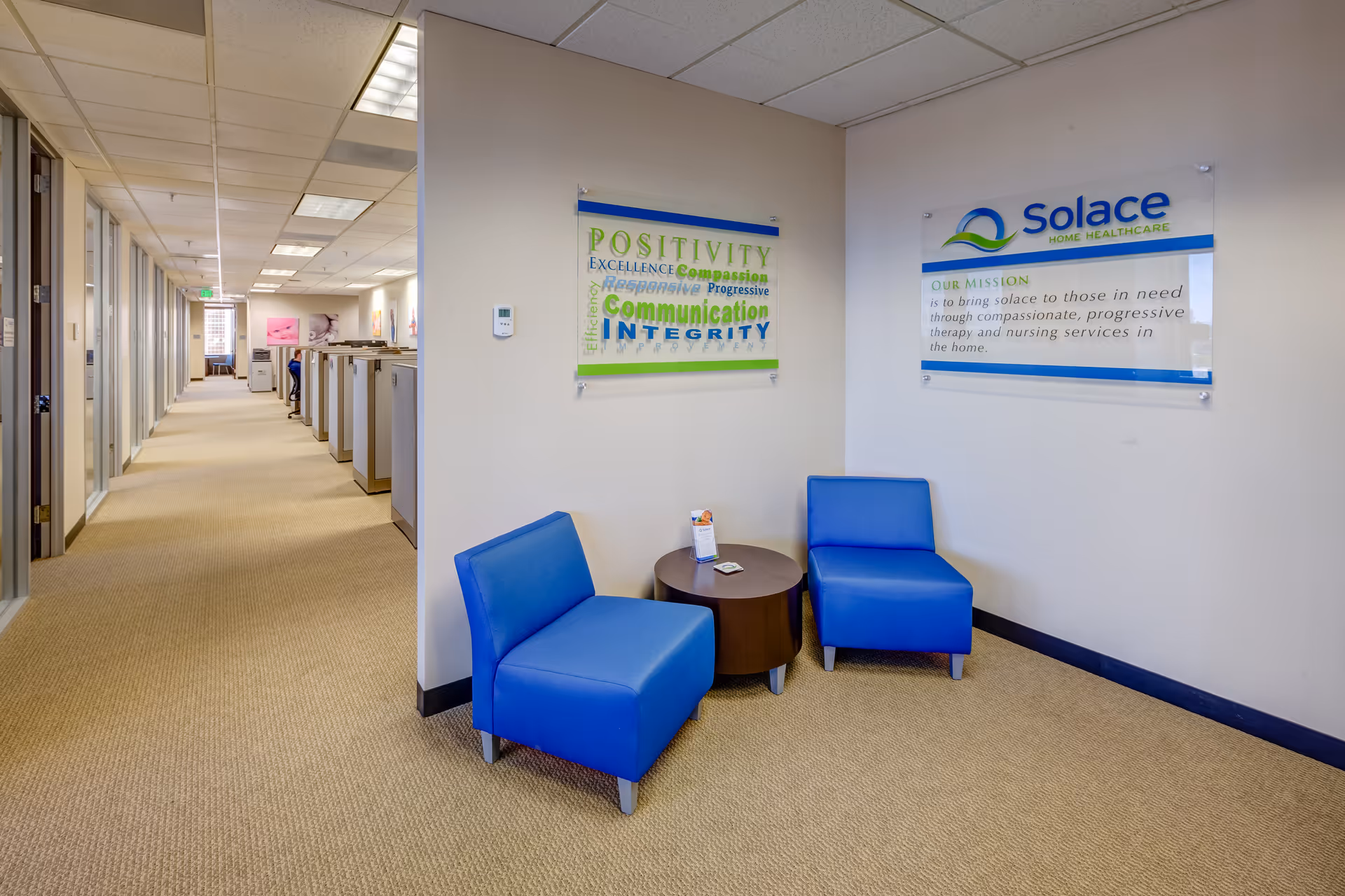 Long interior hallway with beige carpet, blue chairs around a round table, and wall posters.