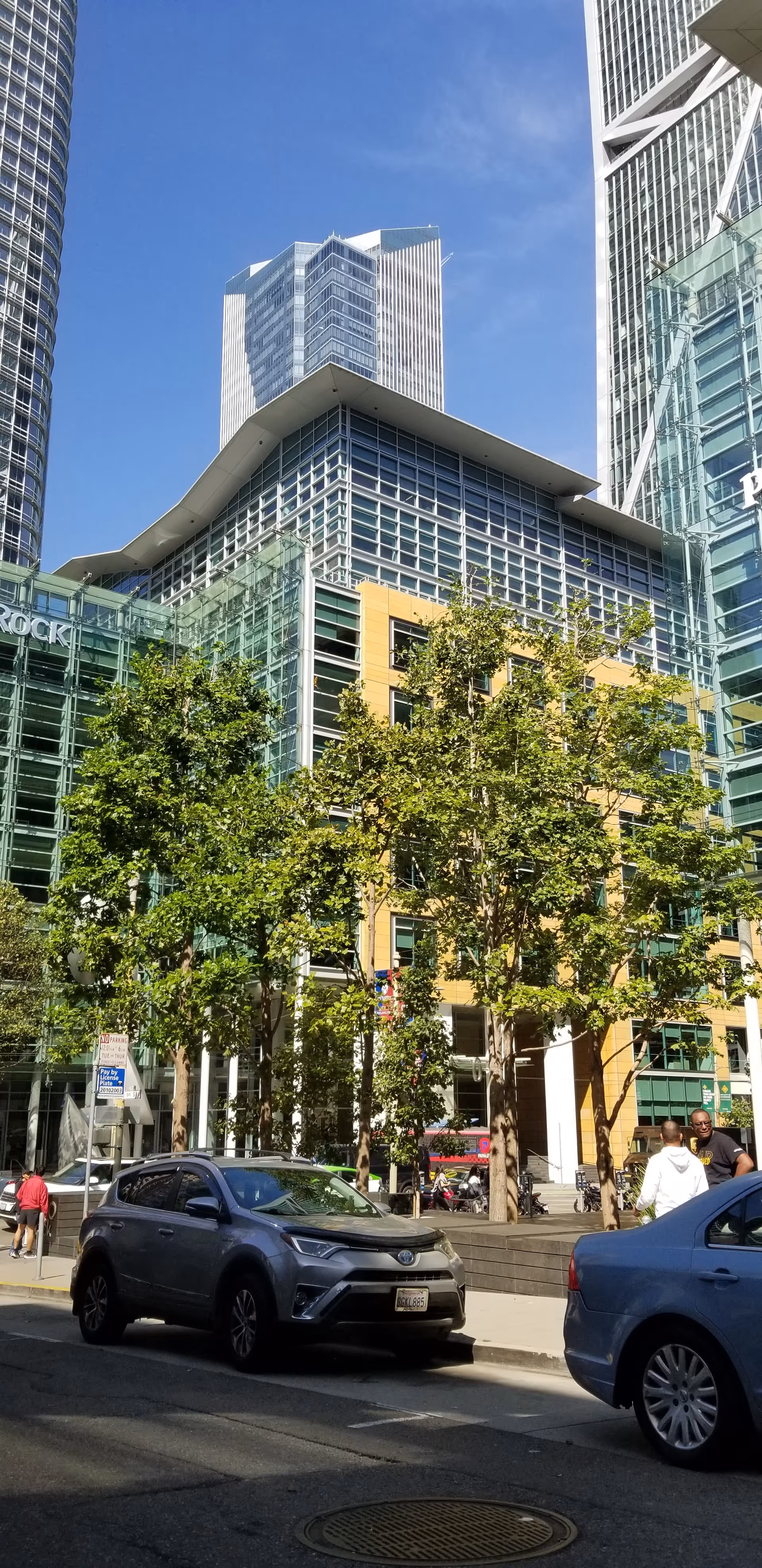 Tall glass skyscrapers surround a tree-lined street with parked cars.