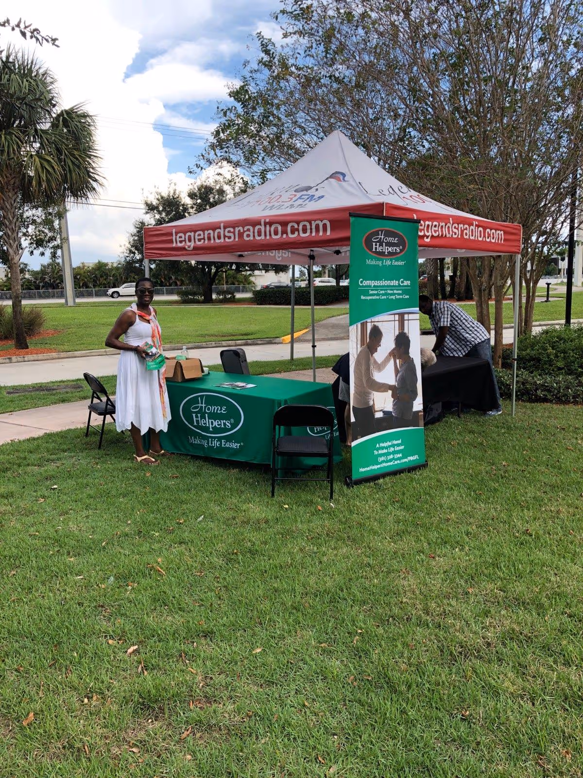 Outdoor Home Helpers booth with a green table and a woman standing on the grass.