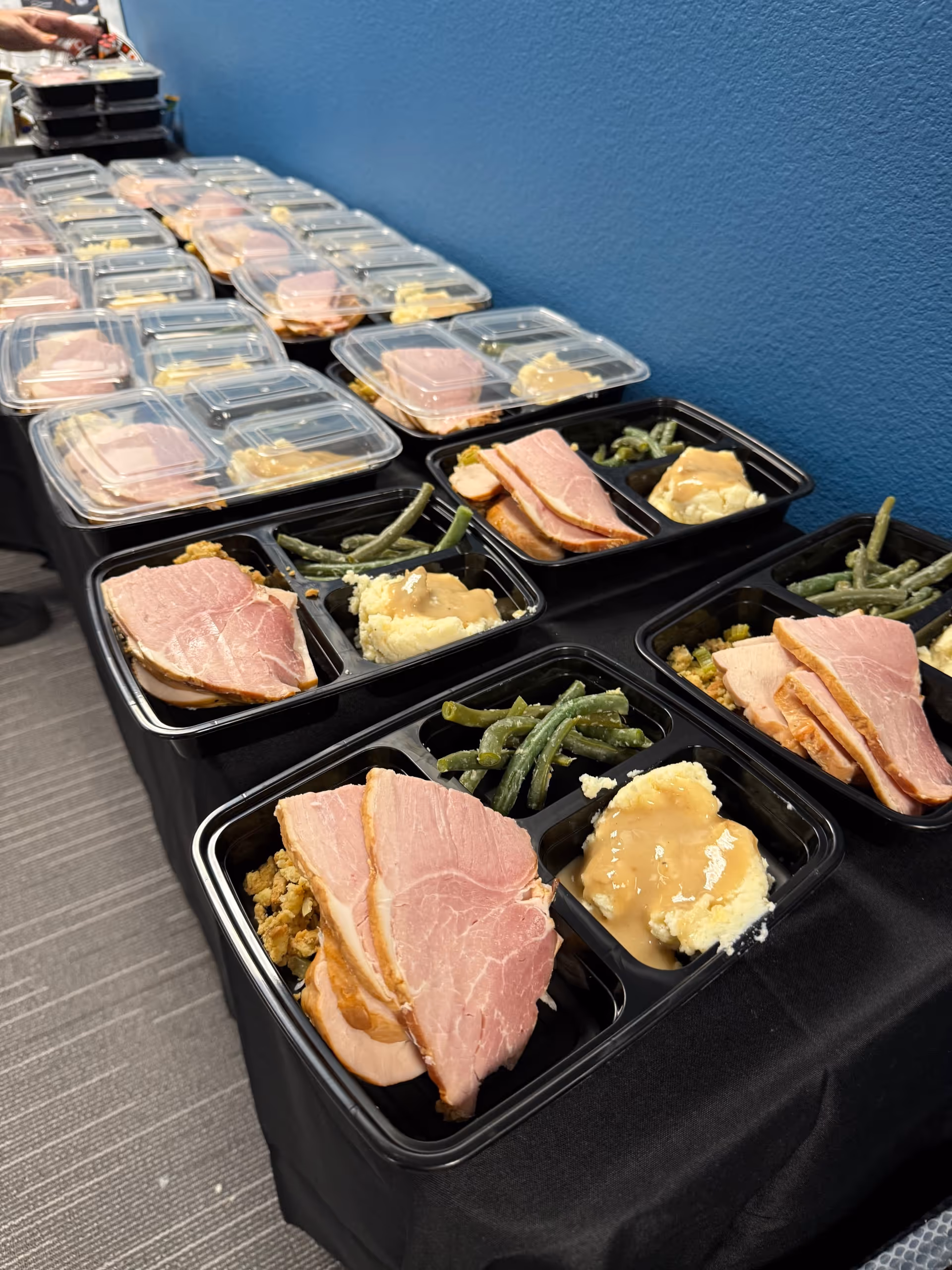 Rows of ready-to-eat meal trays on a table, featuring ham, green beans, and mashed potatoes.