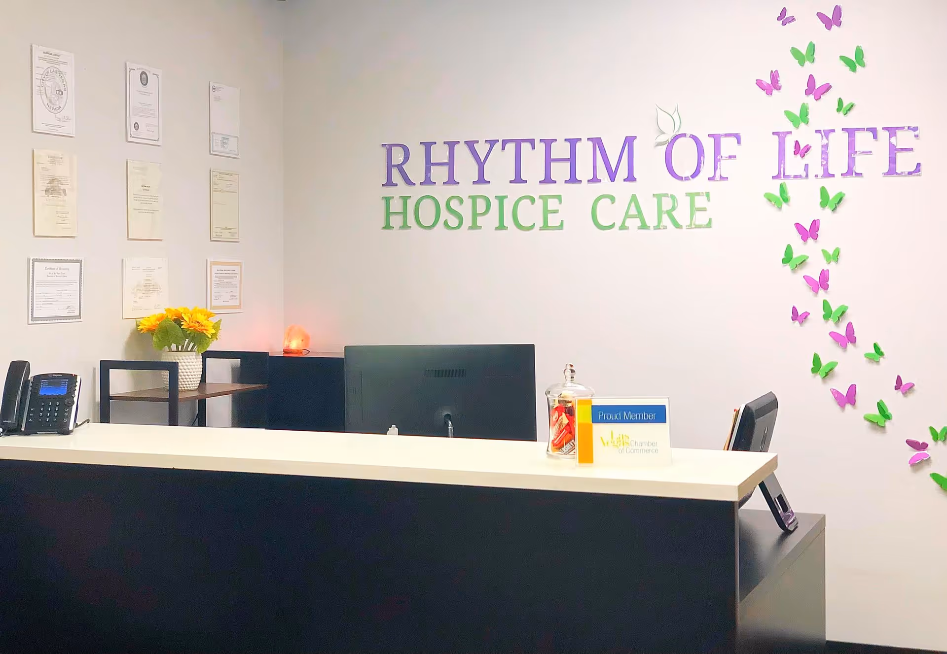 Reception desk area with white counter, computer, flowers, and 'Rhythm of Life Hospice Care' sign.