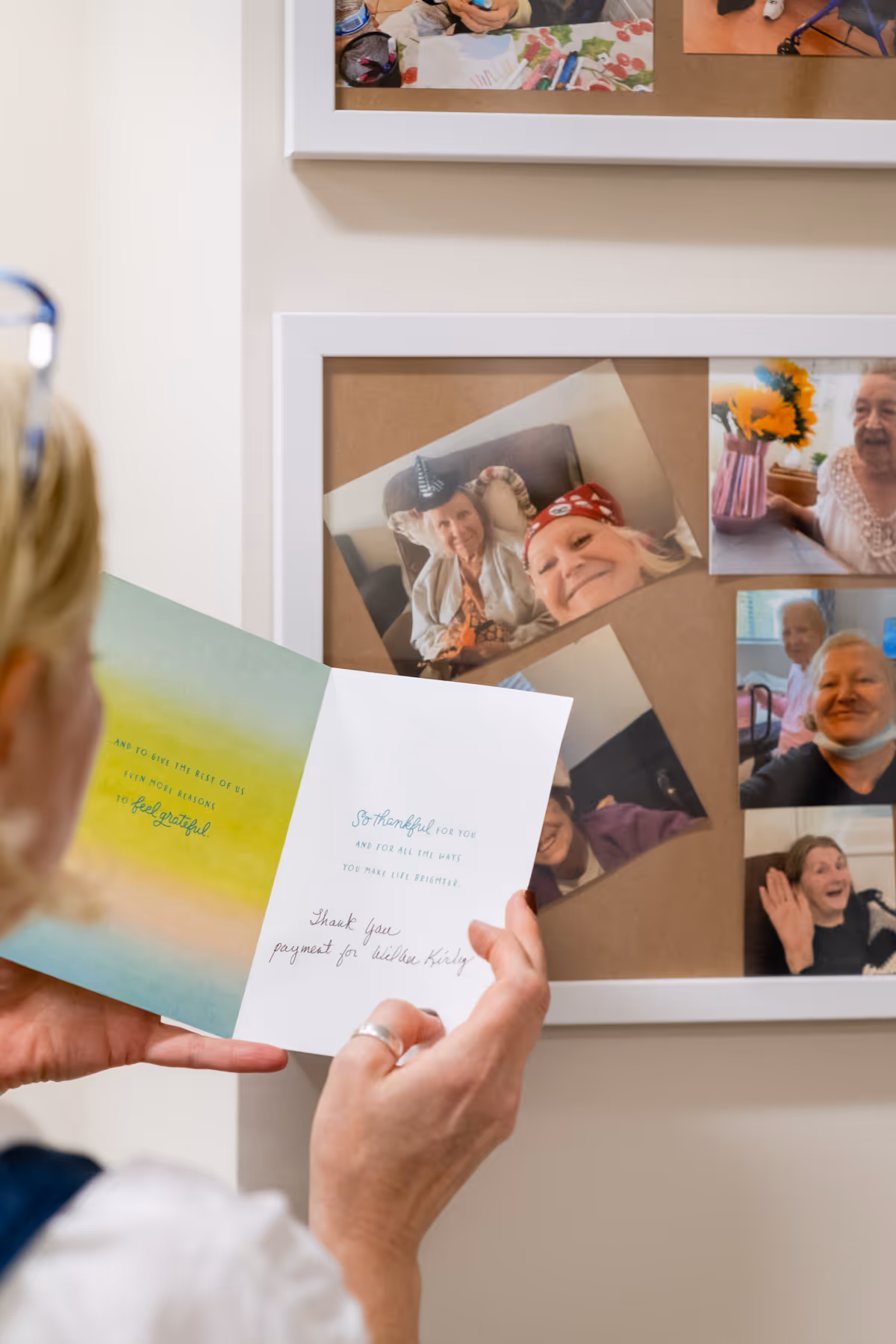 A person holds a greeting card in front of a corkboard collage of smiling elderly photos.