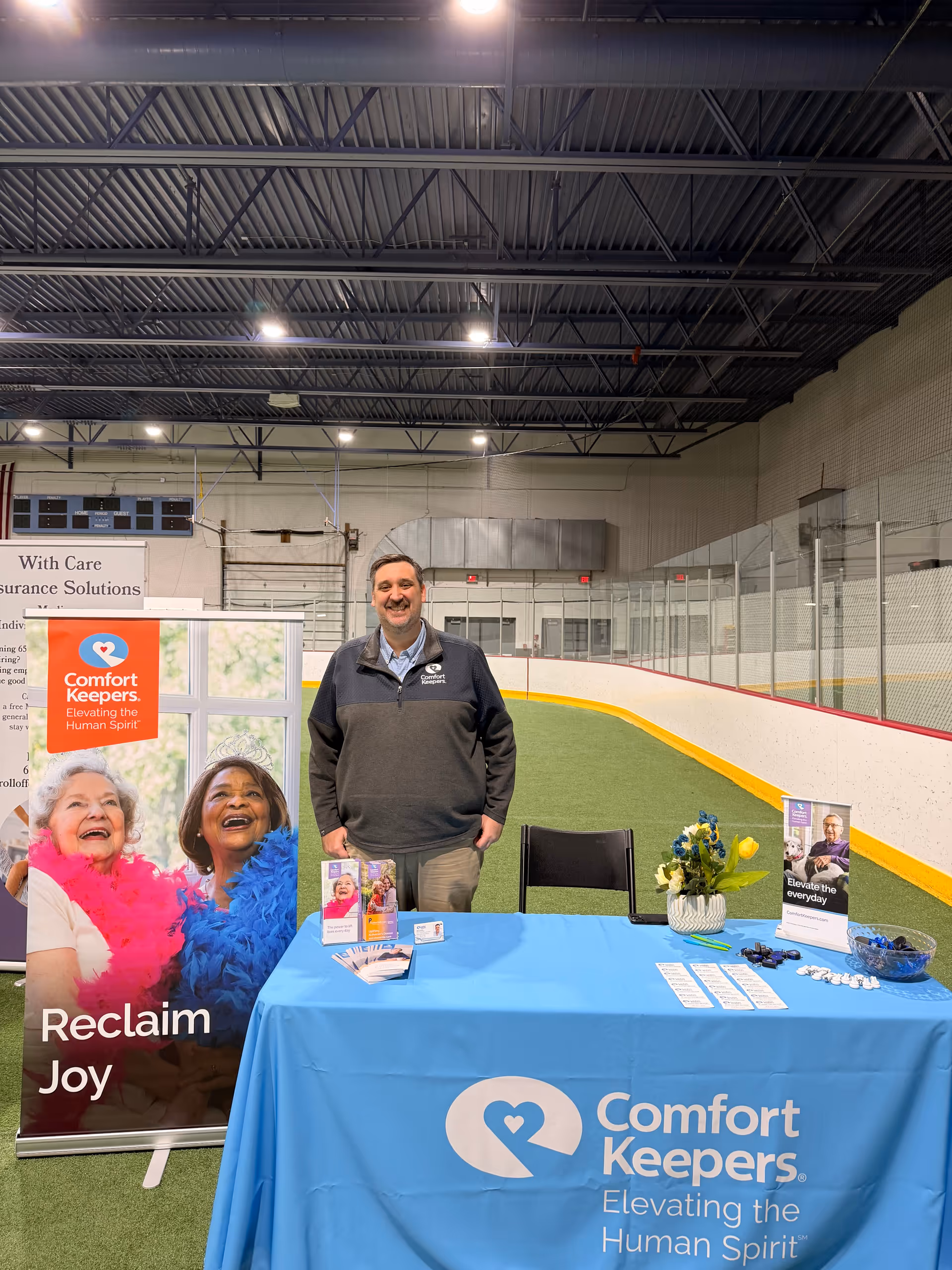 Man stands behind a Comfort Keepers promotional table in an indoor facility with rink boards.