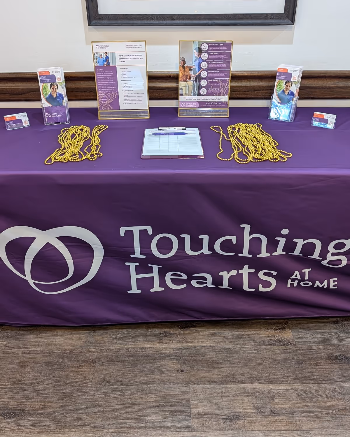 Promotional table for Touching Hearts At Home with brochures and yellow beaded necklaces on purple tablecloth.