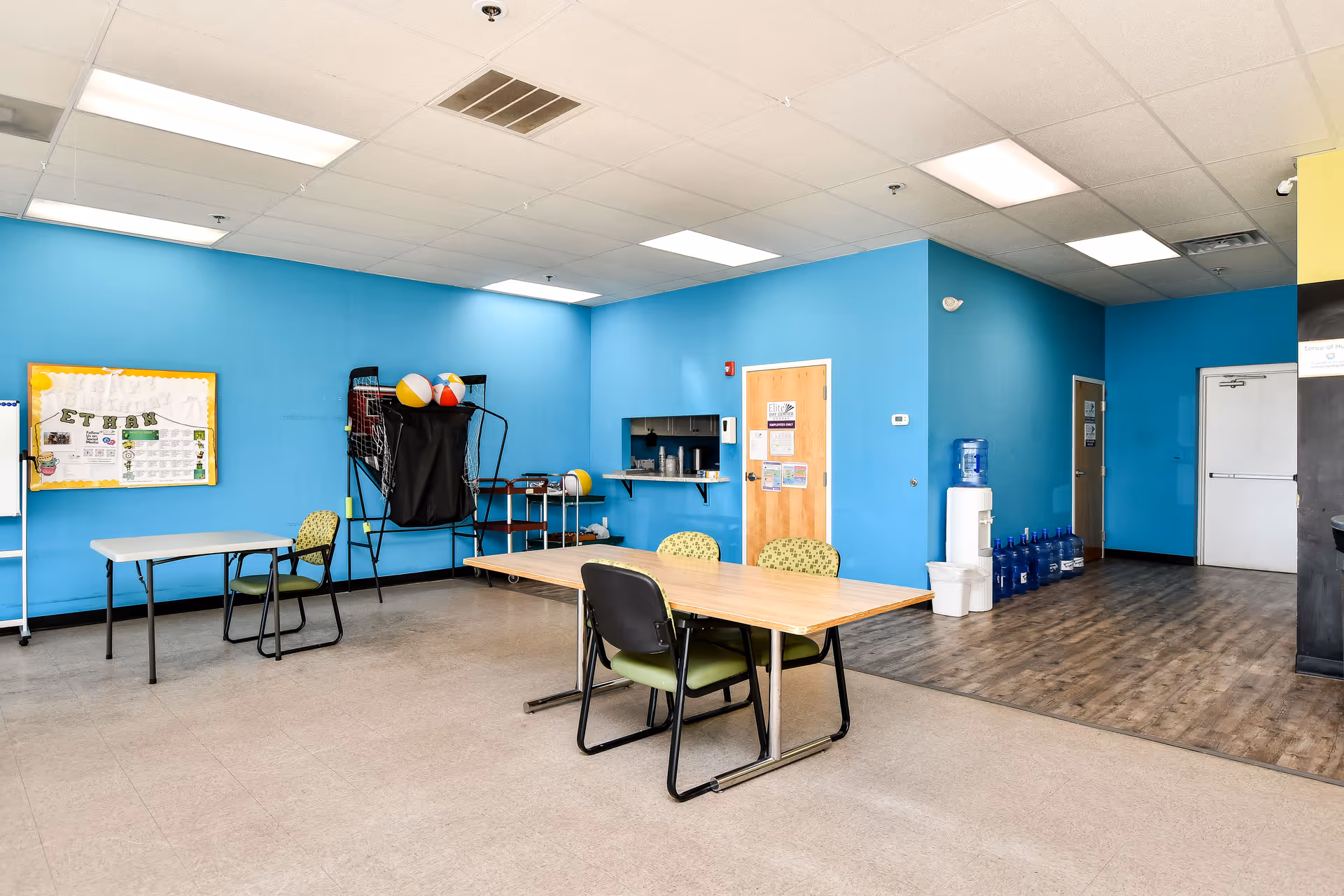 Bright blue walls surround a multipurpose room with tables, chairs, a water cooler, and exercise equipment.